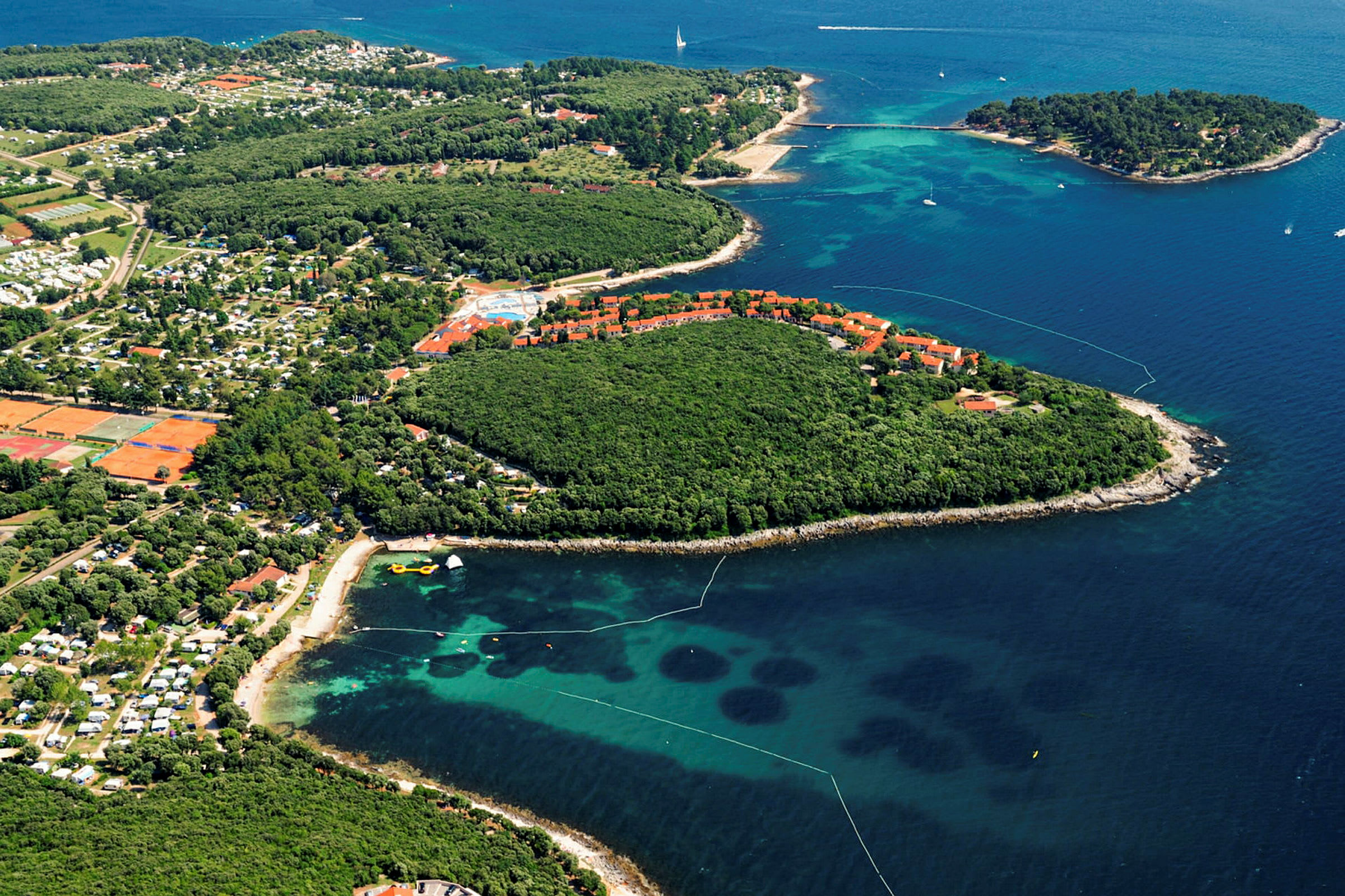 Flygbild över en kuststräcka med skog, byggnader i rött tak, campingområden och havet. (AI-genererad text)