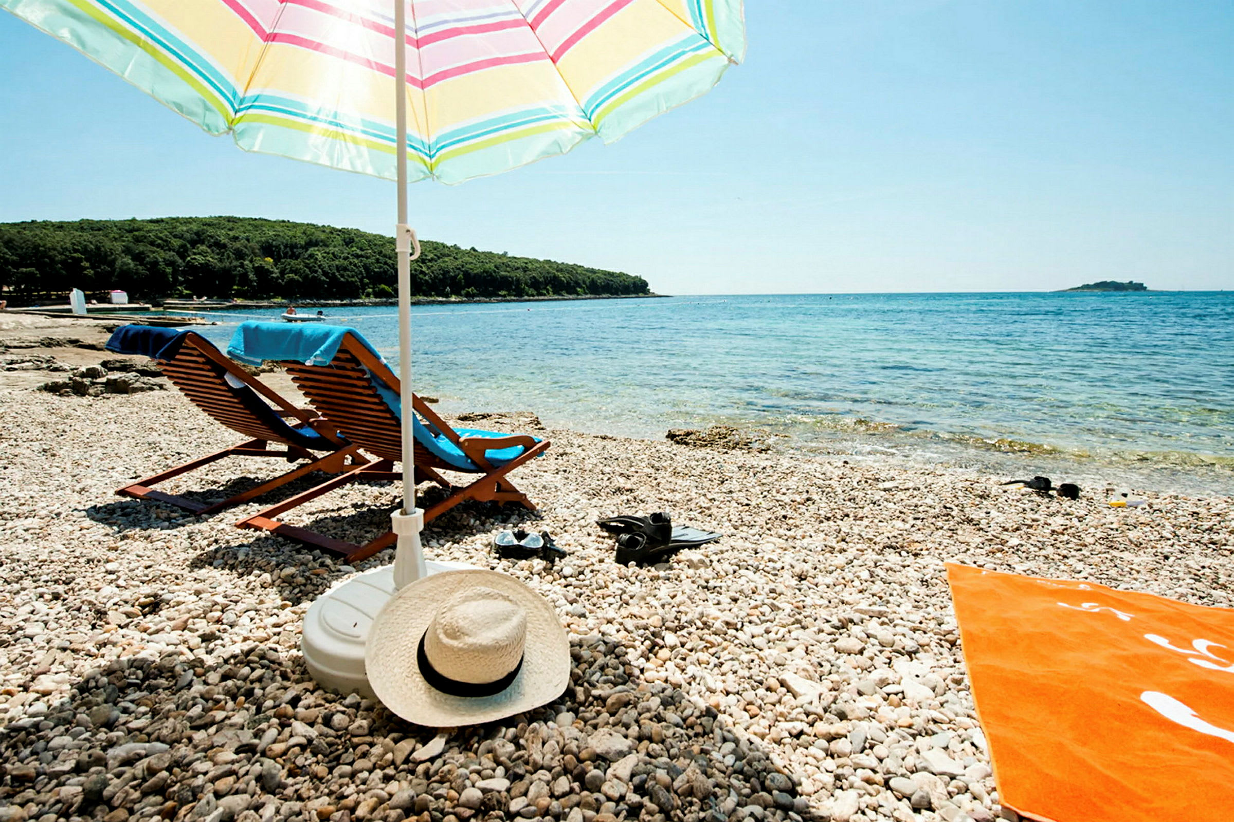 En stenstrand med två solstolar under ett randigt parasoll. Det turkosa havet syns i bakgrunden. (AI-genererad text)