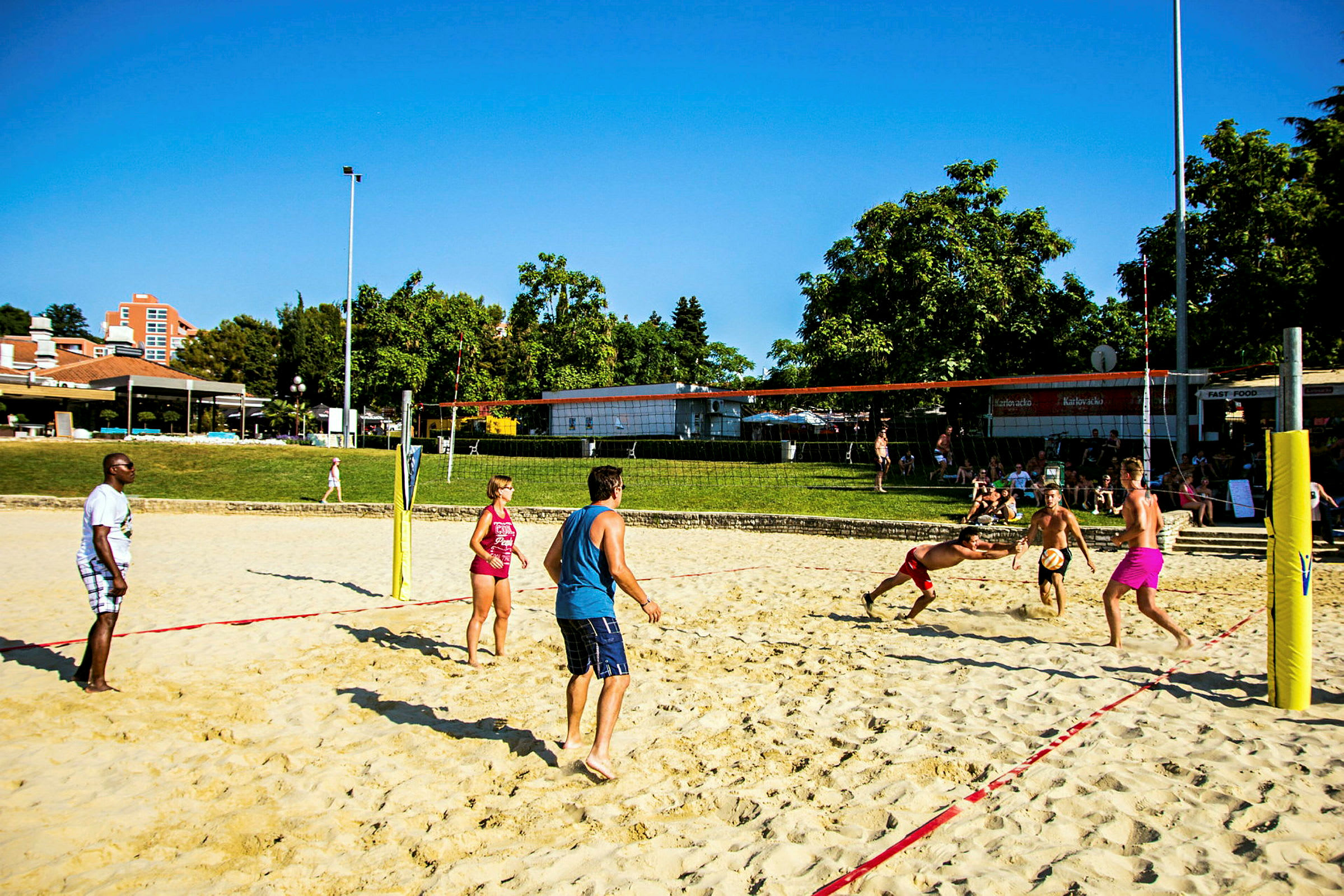 Människor spelar beachvolleyboll på en sandplan i ett utomhusområde med träd och byggnader i bakgrunden. (AI-genererad text)