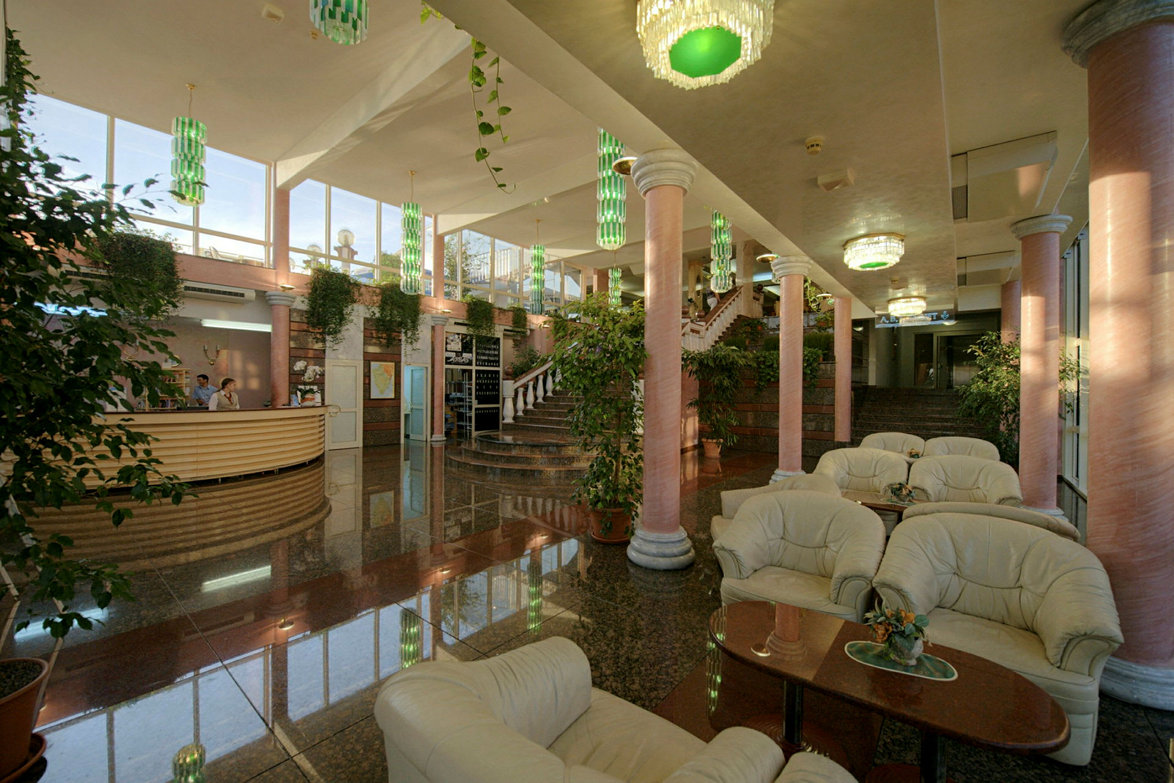 Interiör av en hotellobby med marmorgolv, pelare i rosa ton, belysning med gröna ljuskronor, och sittgrupper med vita möbler. (AI-genererad text)