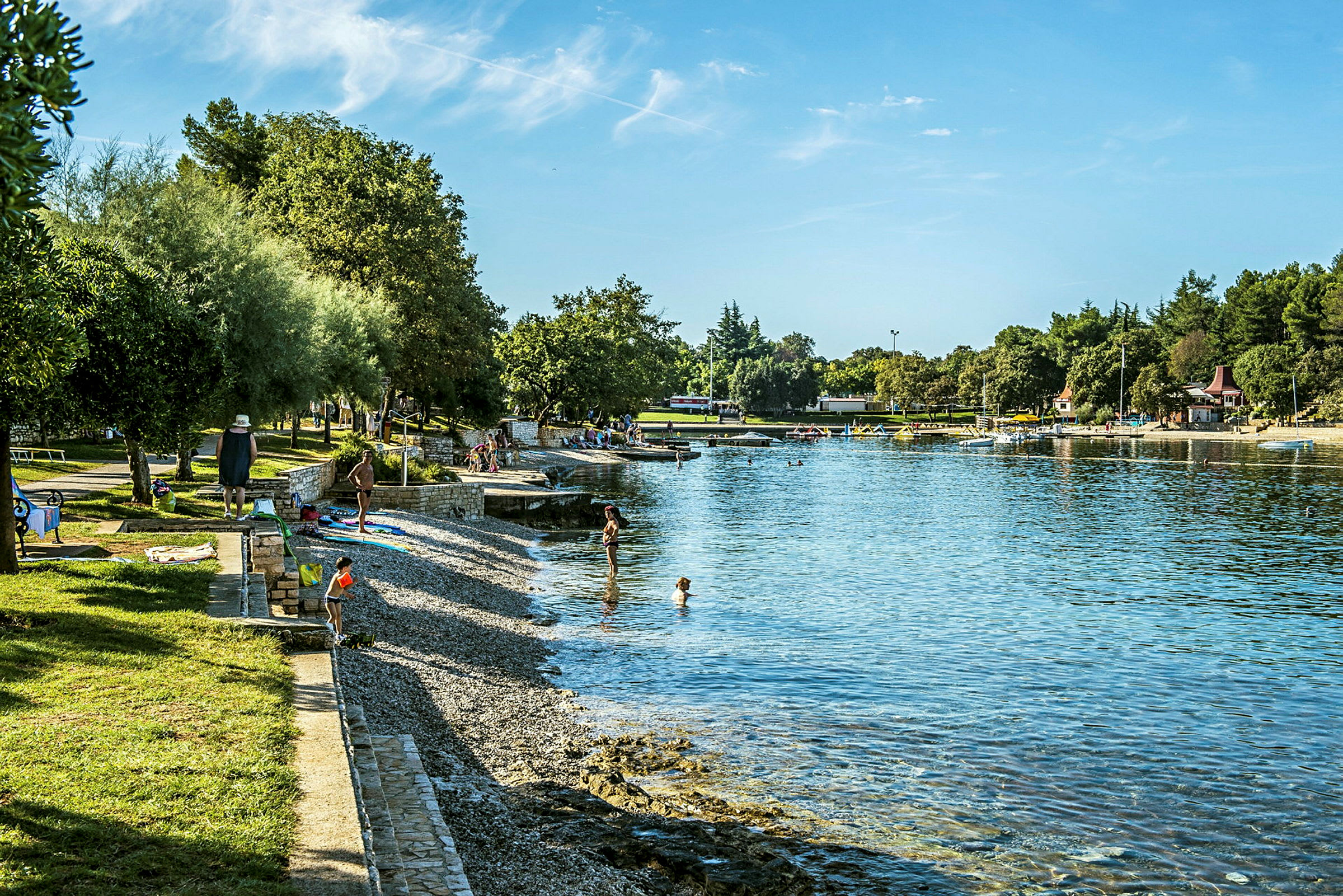 Bild på en strand med klappersten vid en lugn vik. Flera personer syns vid vattenbrynet och på gräsmatta bredvid strandpromenaden. (AI-genererad text)