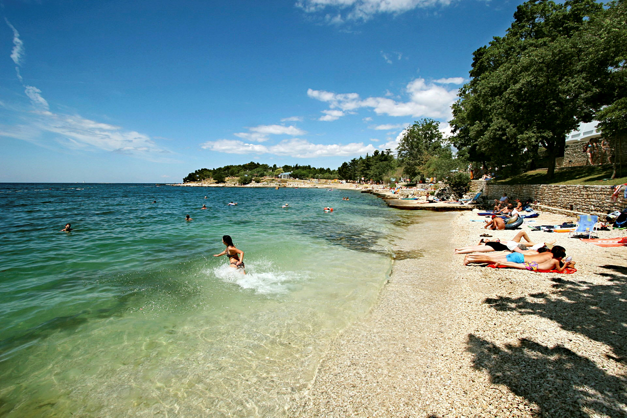 En strand med småsten där människor solar och badar i det klarblå havet. (AI-genererad text)