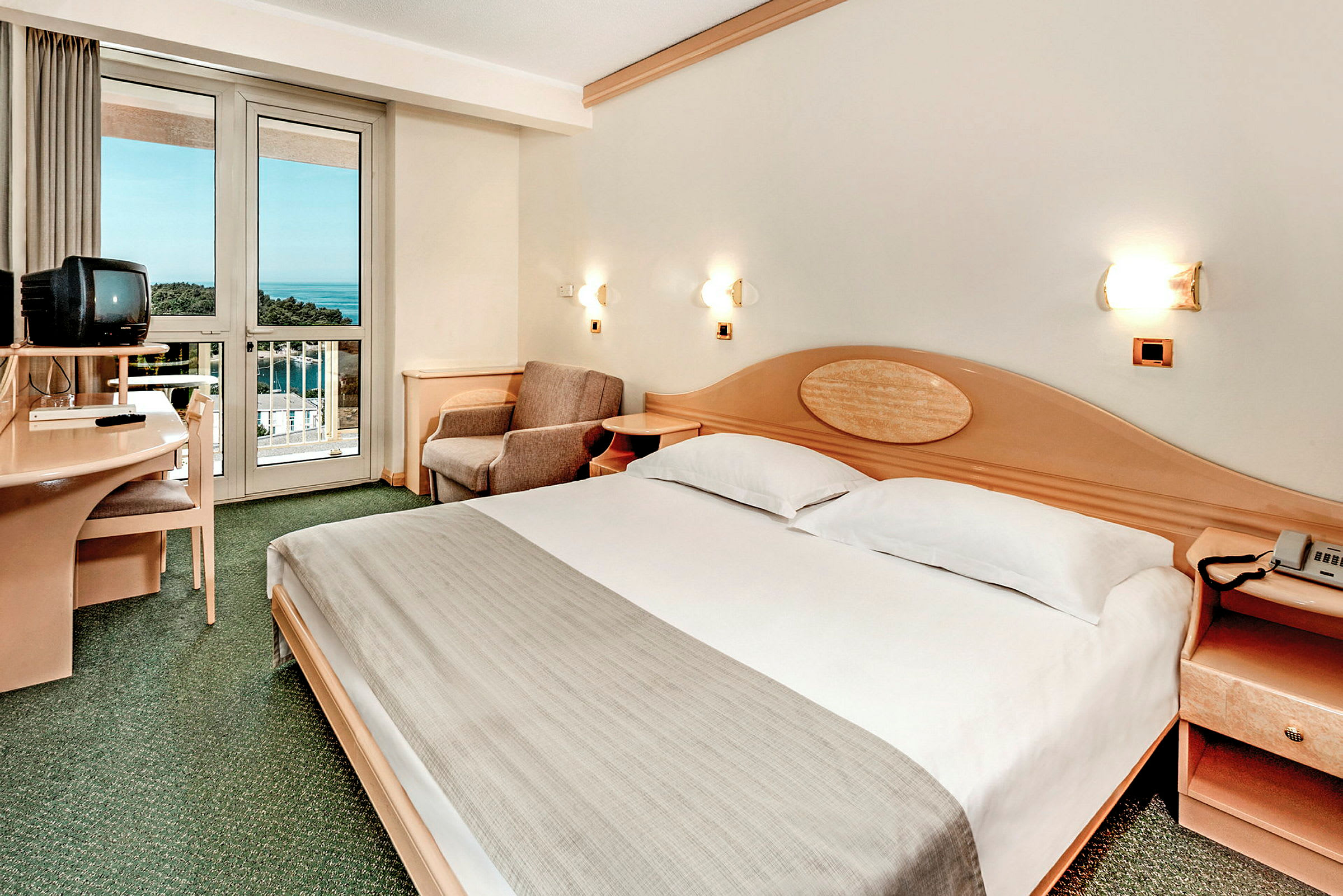Ett hotellrum med en dubbelsäng, en TV på ett skrivbord och en balkong med utsikt över havet. (AI-genererad text)