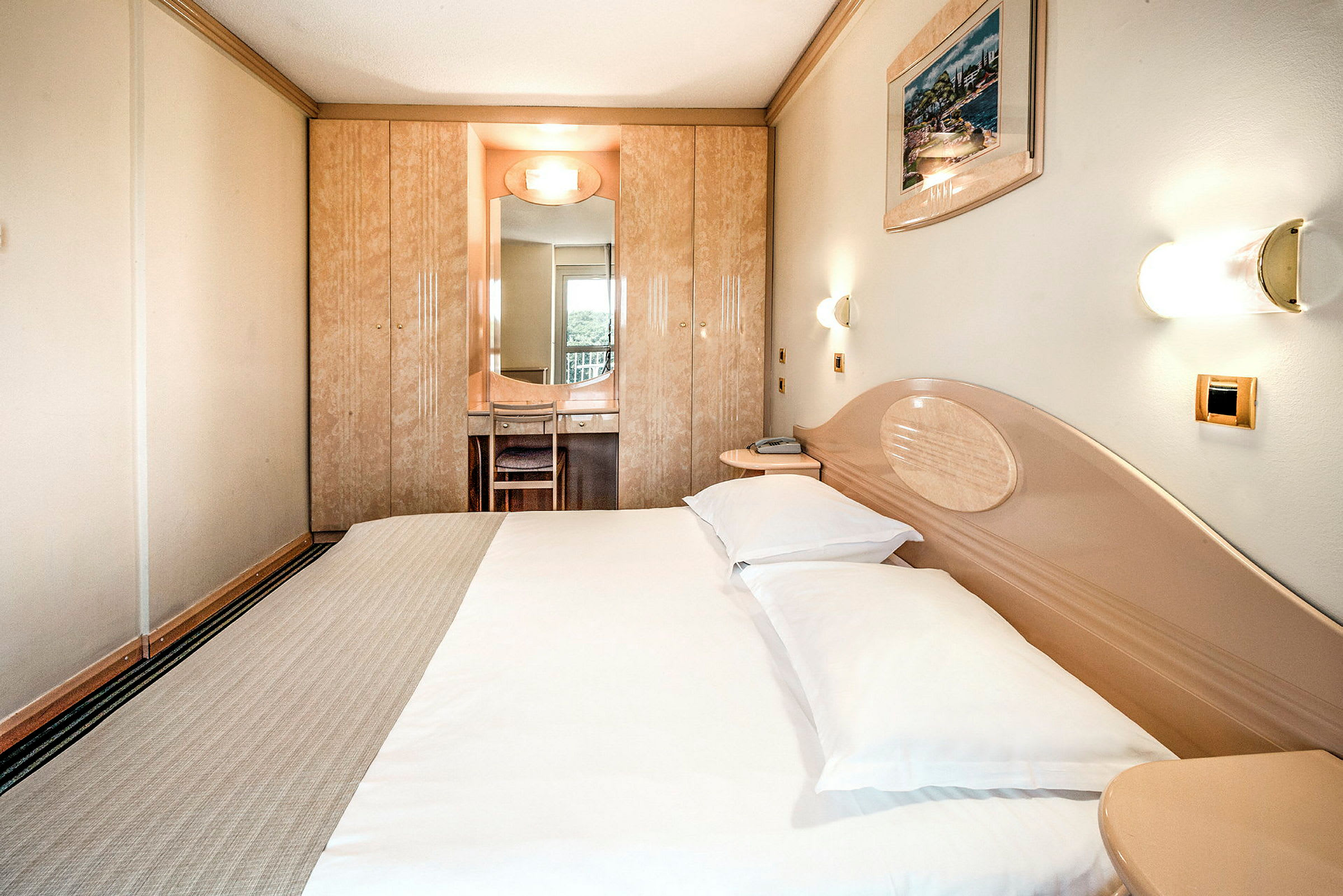 Ett hotellrum med en dubbelsäng, väggmonterade lampor och en inbyggd garderob med en spegel framför en stol. (AI-genererad text)