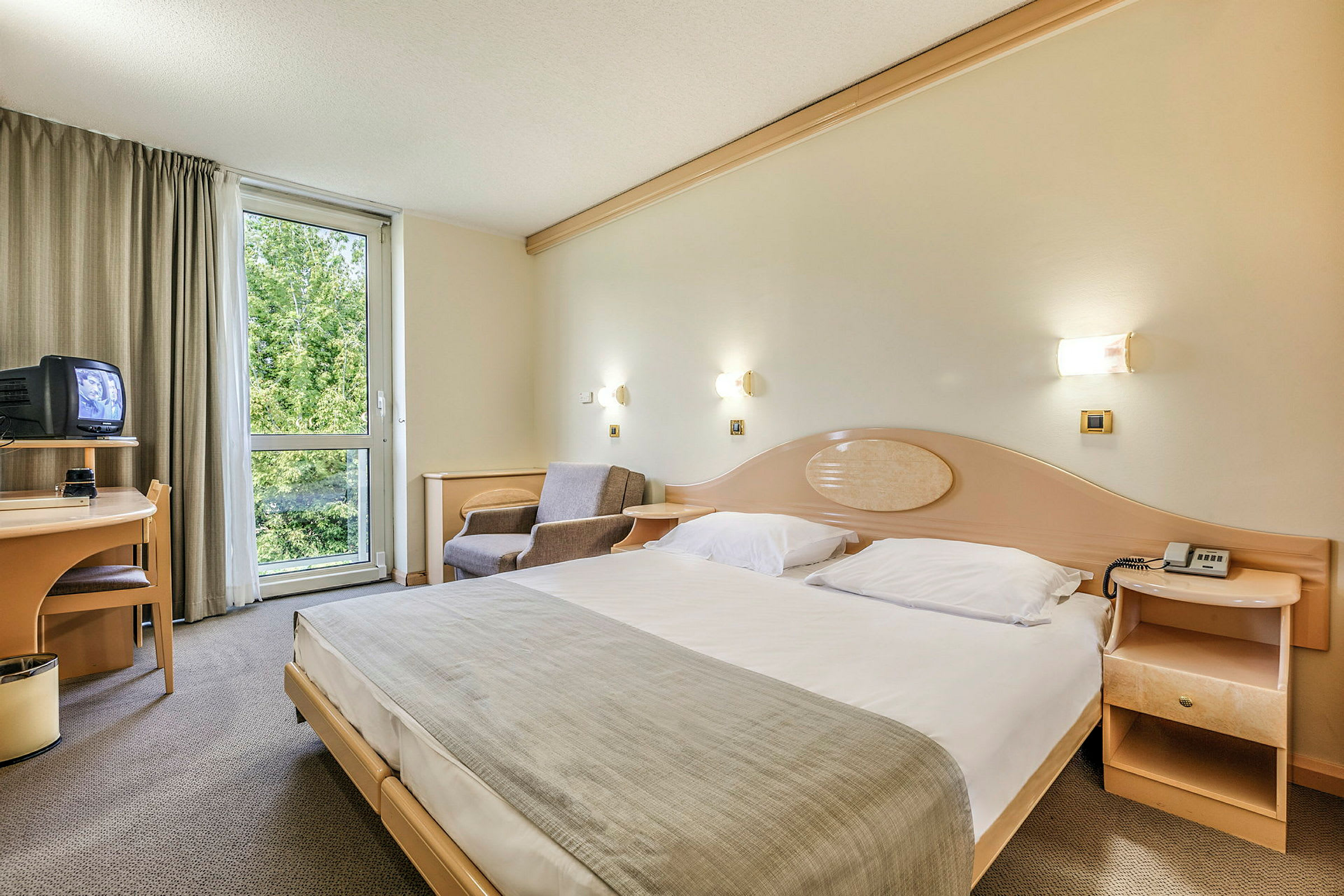 Hotelrum med en stor säng, en fåtölj, ett skrivbord med en TV och ett fönster med utsikt över grönska. (AI-genererad text)