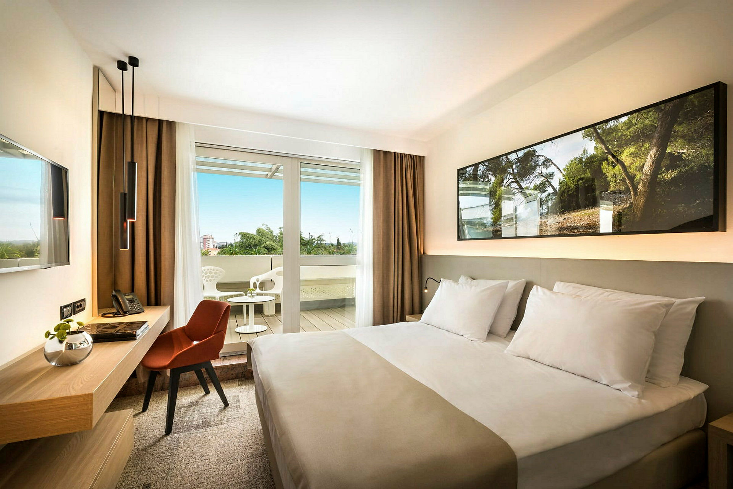 Hotellrum med en säng, skrivbord och stol, samt en balkong med utemöbler. (AI-genererad text)