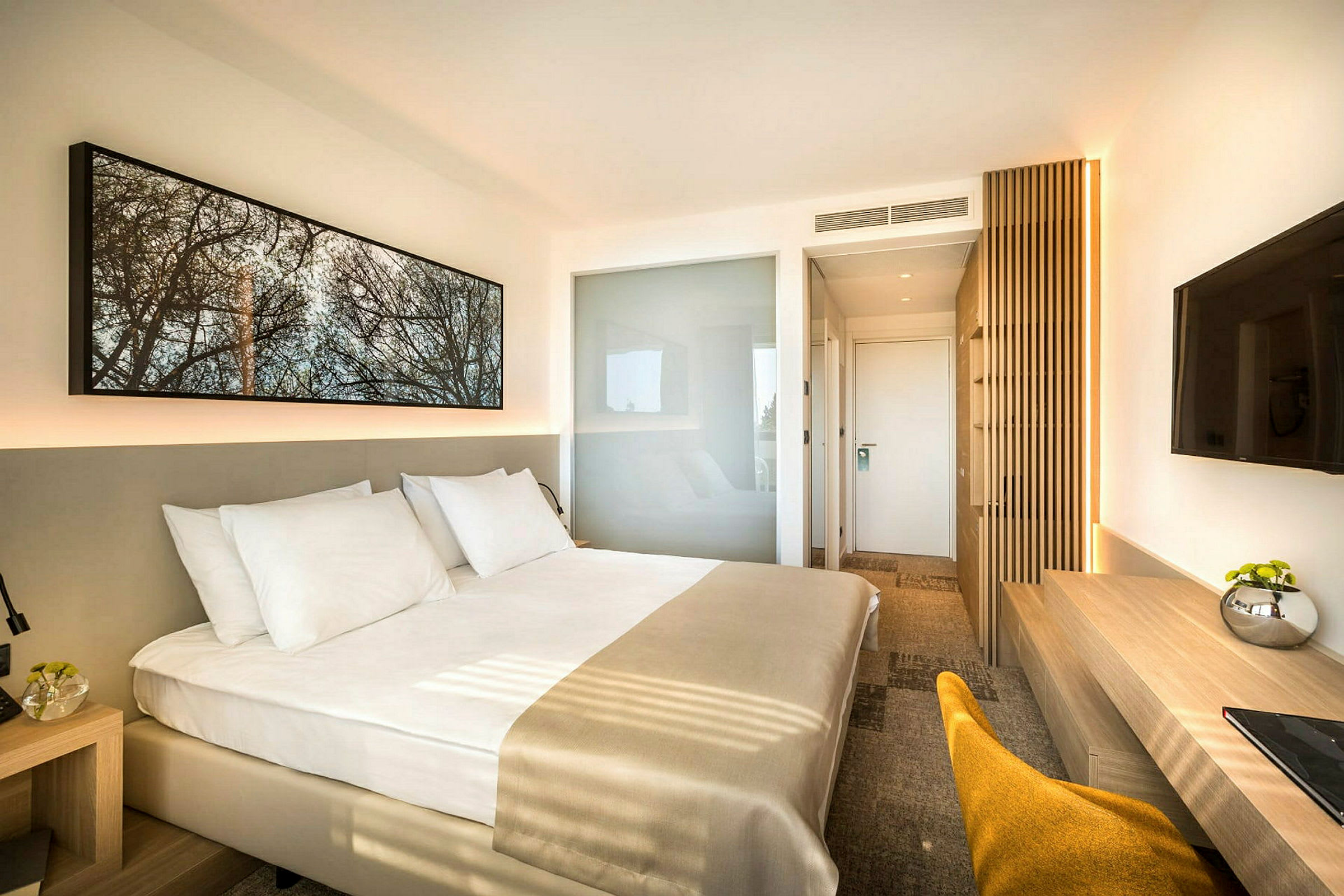 Ett modernt hotellrum med en stor säng klädd i vita sängkläder. Rummet har en minimalistisk inredning med en väggmonterad TV, ett skrivbord och en stol. (AI-genererad text)