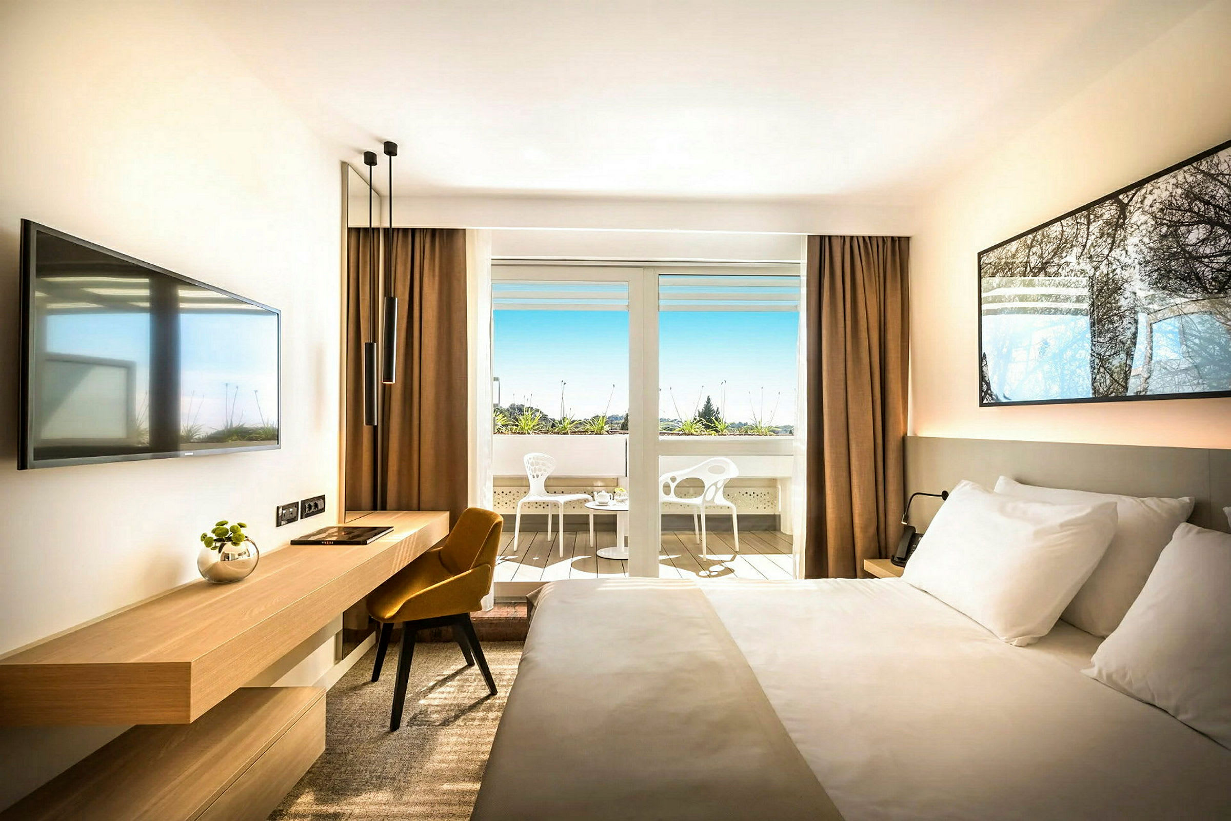 Ett hotellrum med en stor säng, en skrivbordsmöbel, en väggmonterad TV, och stora glasdörrar som leder ut till en terrass med stolar samt utsikt mot grönska. (AI-genererad text)