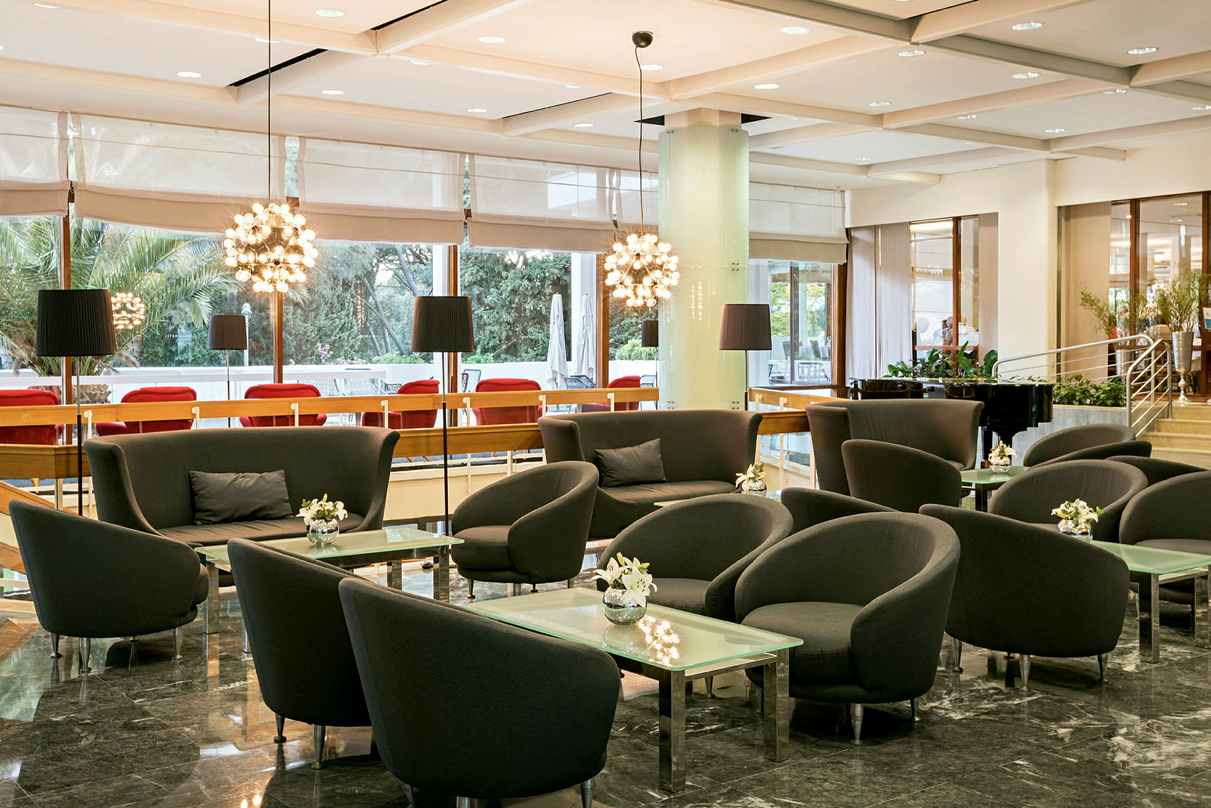 En välbelyst lounge med sittgrupper bestående av fåtöljer och soffor runt glasbord, omgivna av stora fönster och modern inredning. (AI-genererad text)