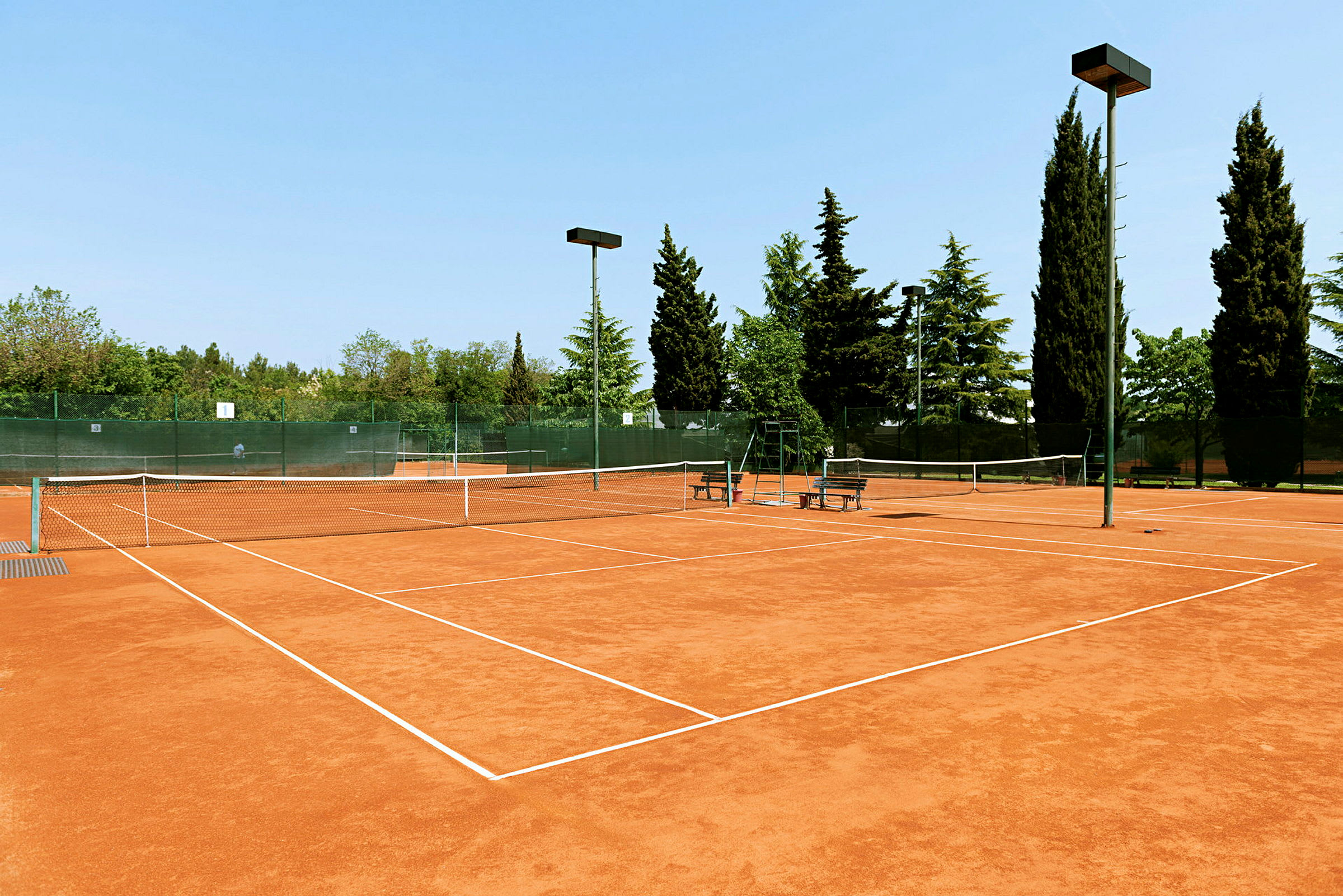 En tennisplan med grusunderlag omgiven av träd och stängsel. (AI-genererad text)