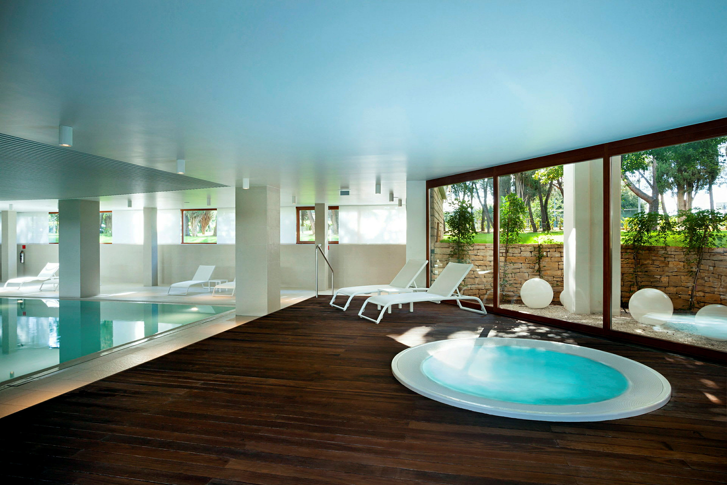 Inomhusbassäng med solstolar och jacuzzi bredvid, samt glasvägg med utsikt mot en grönskande trädgård. (AI-genererad text)