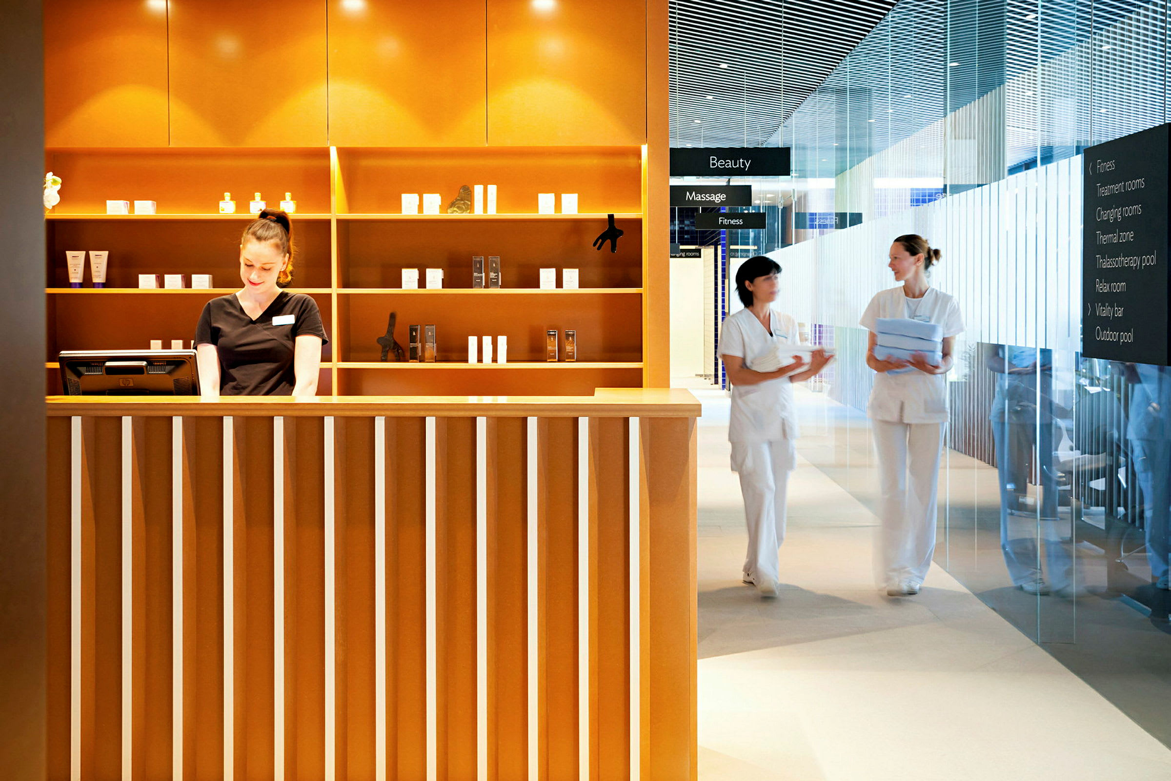 Reception av spa- eller skönhetsanläggning med hyllor fyllda med produkter och två personer i arbetskläder samtalet vid sidan om. (AI-genererad text)