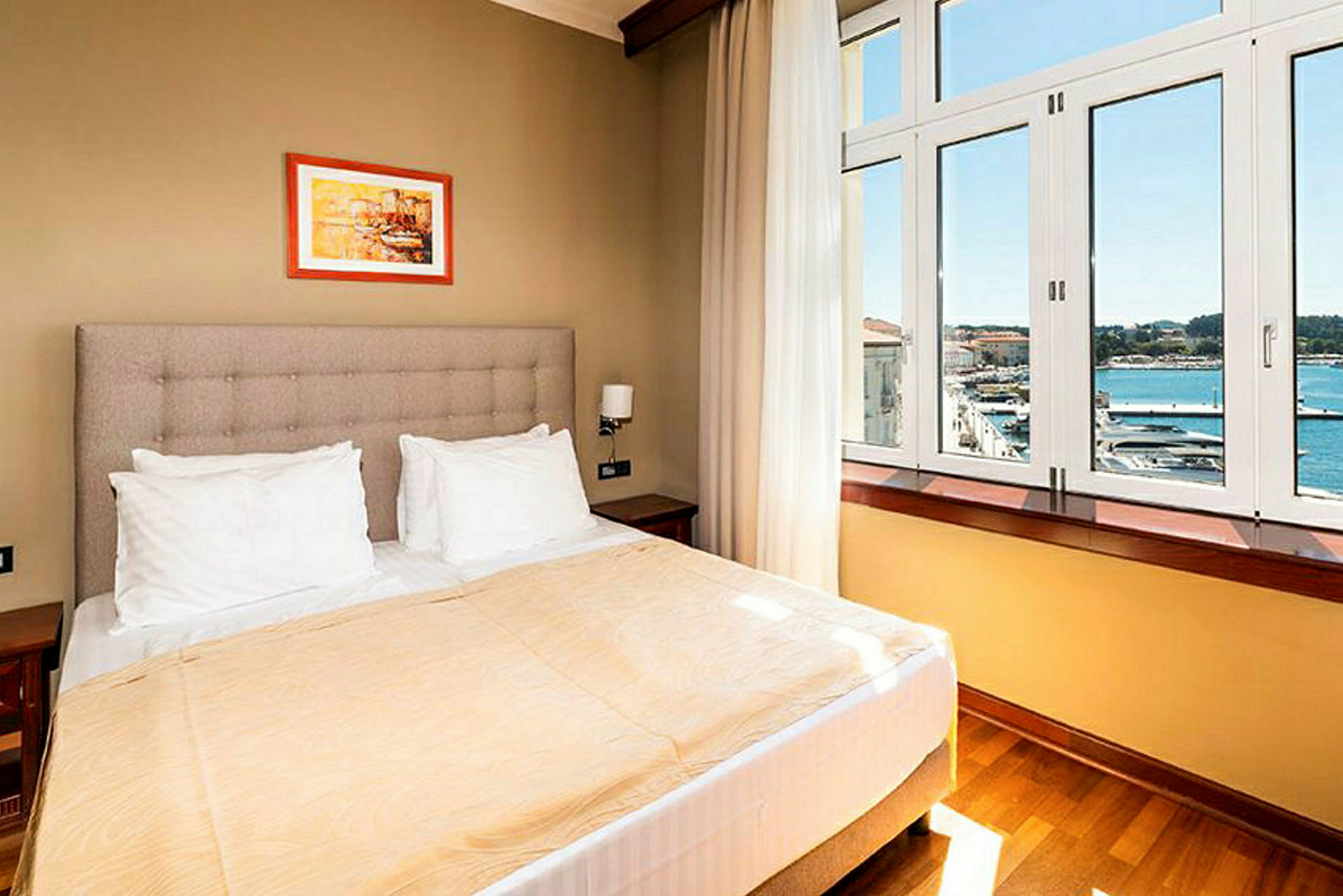 Ett hotellrum med en dubbelsäng, vita kuddar, beige överkast och ett stort fönster som vetter mot en havsutsikt. (AI-genererad text)