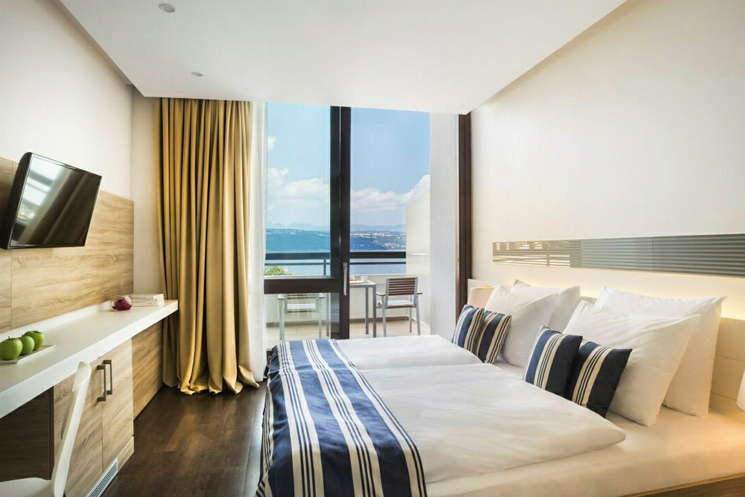 Ett hotellrum med en dubbelsäng, sängkläder i blå- och vitrandigt mönster, och en stor glasvägg som visar en balkong med utsikt över havet. Rummet har även en TV och skrivbord. (AI-genererad text)