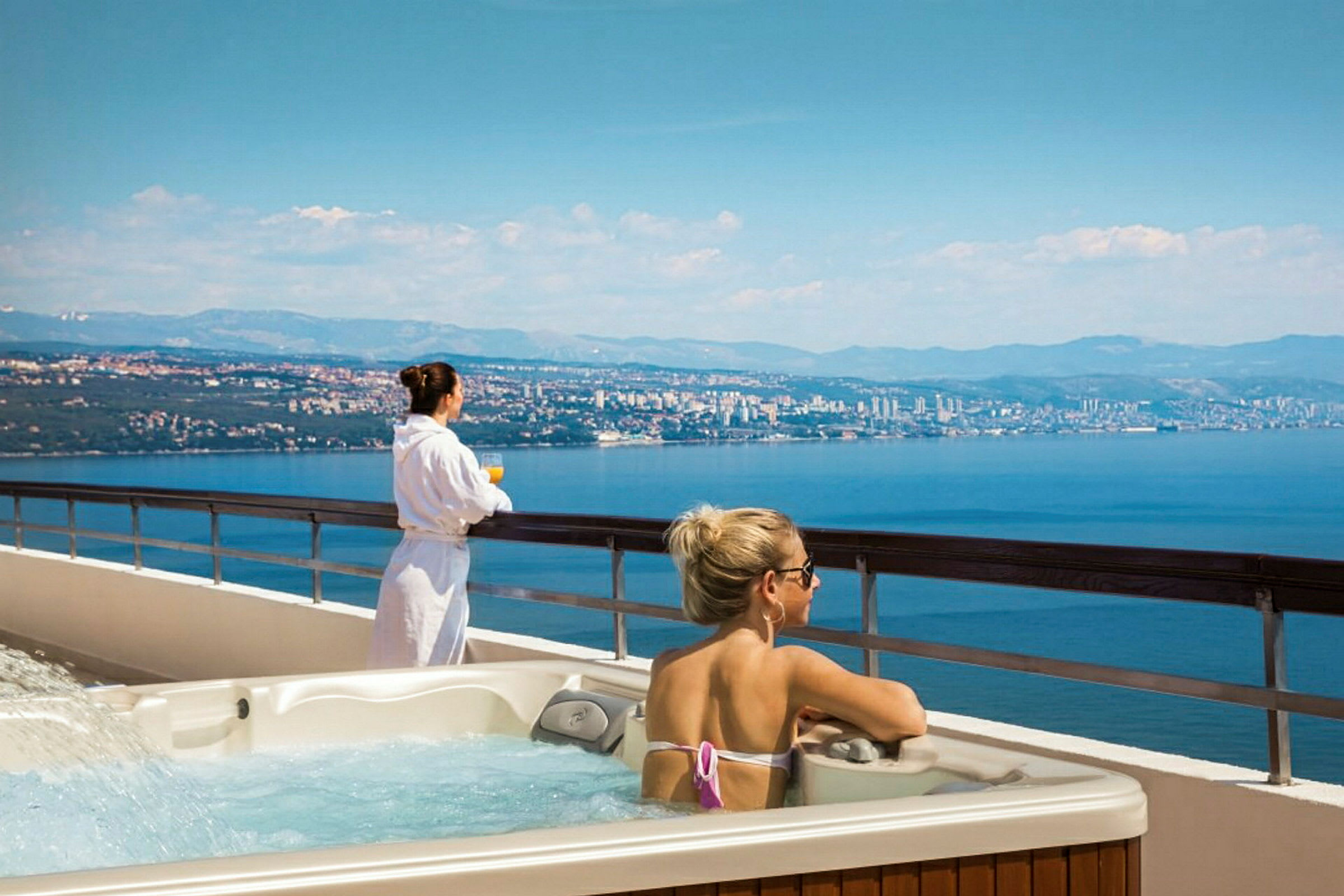 Personer njuter av utsikten och relaxar vid en Jacuzzi på en balkong med panoramavy över havet och en stad. (AI-genererad text)
