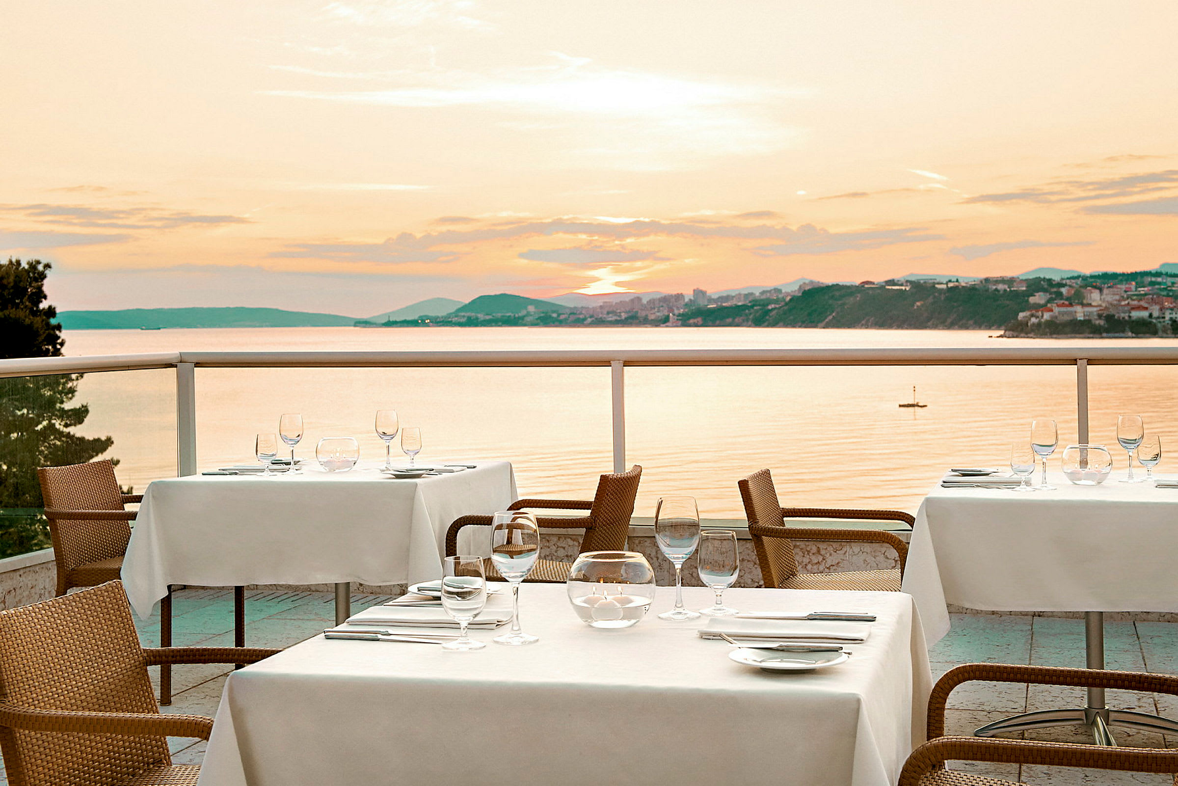 Bord med vita dukar på en terrass med utsikt över havet och solnedgången. (AI-genererad text)