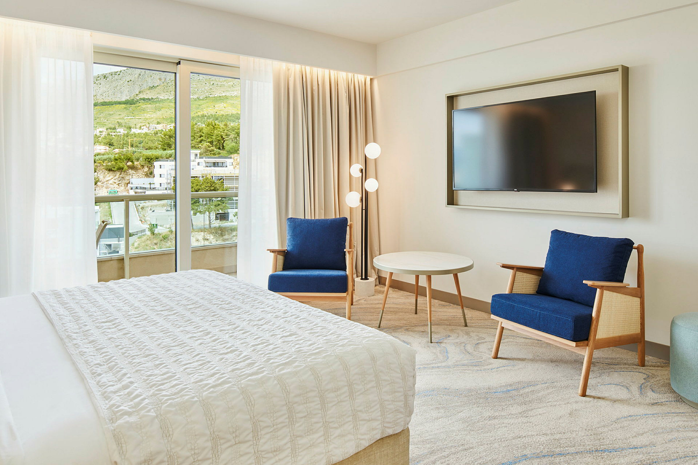 Hotellrum med säng, två blåa fåtöljer, ett runt bord och en väggmonterad TV. Balkong och grön utsikt syns genom glasdörren. (AI-genererad text)
