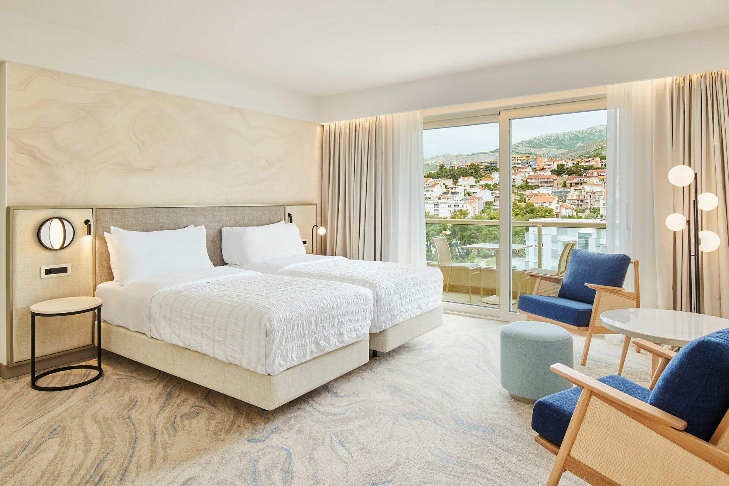 Hotellrum med två enkelsängar, sittgrupp och balkong med utsikt över en stad. (AI-genererad text)