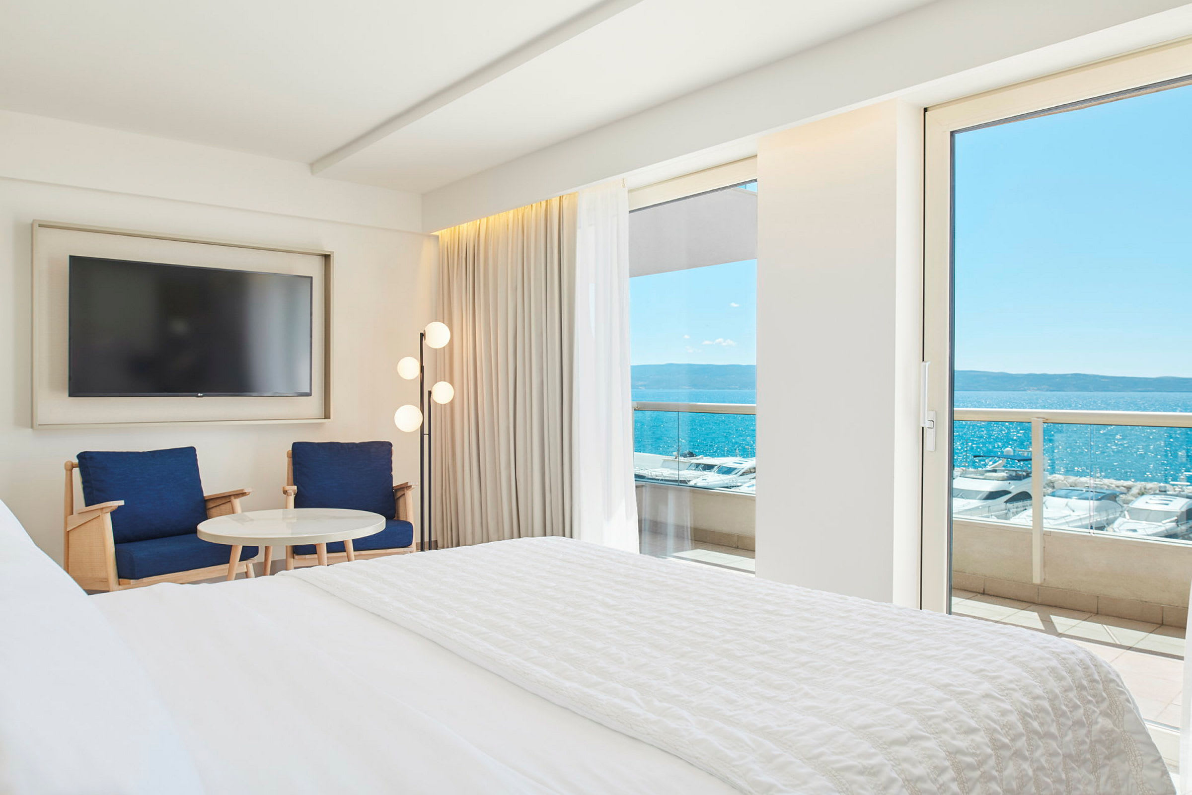 Interiör av ett hotellrum med en stor säng, två stolar, ett bord och en tv. Utsikt över havet genom en balkongdörr. (AI-genererad text)