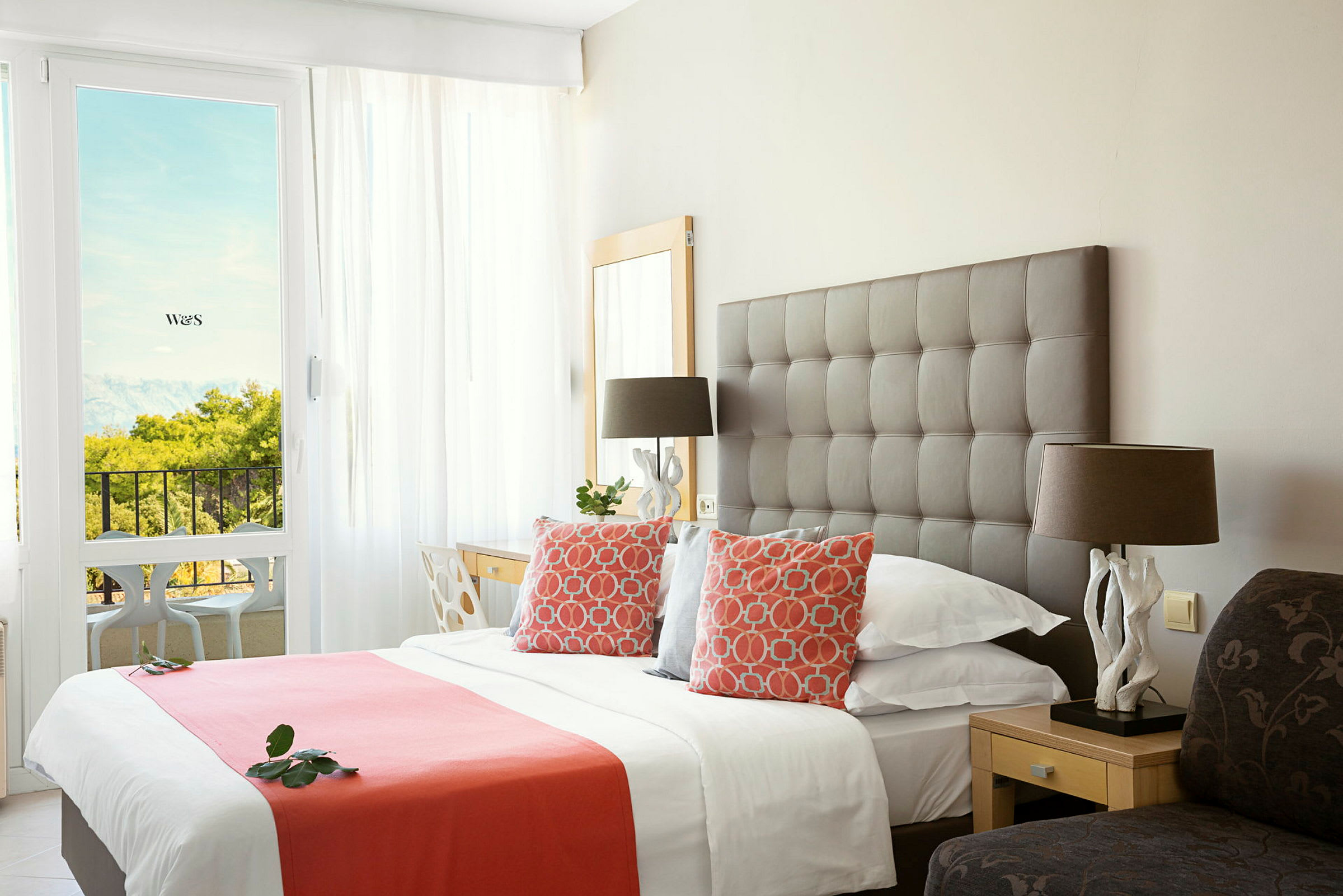 Ett elegant hotellrum med en stor säng, dekorativa kuddar och en solig balkong utsikt. (AI-genererad text)