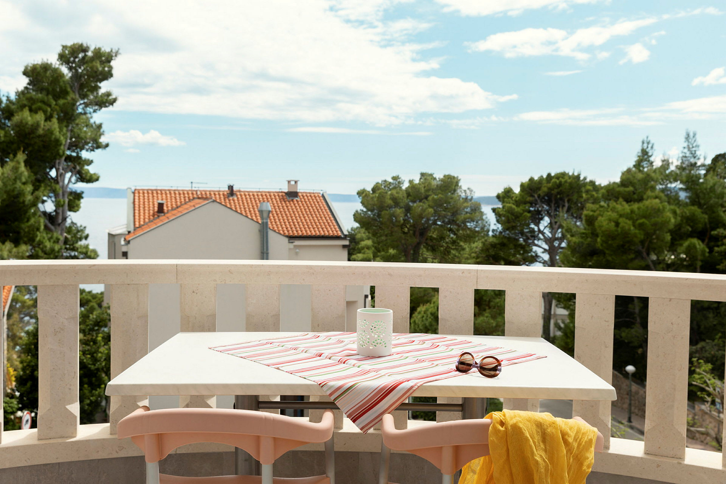 En balkong med bord täckt av randig duk, solglasögon och gul sjal, med utsikt mot havet och hus. (AI-genererad text)