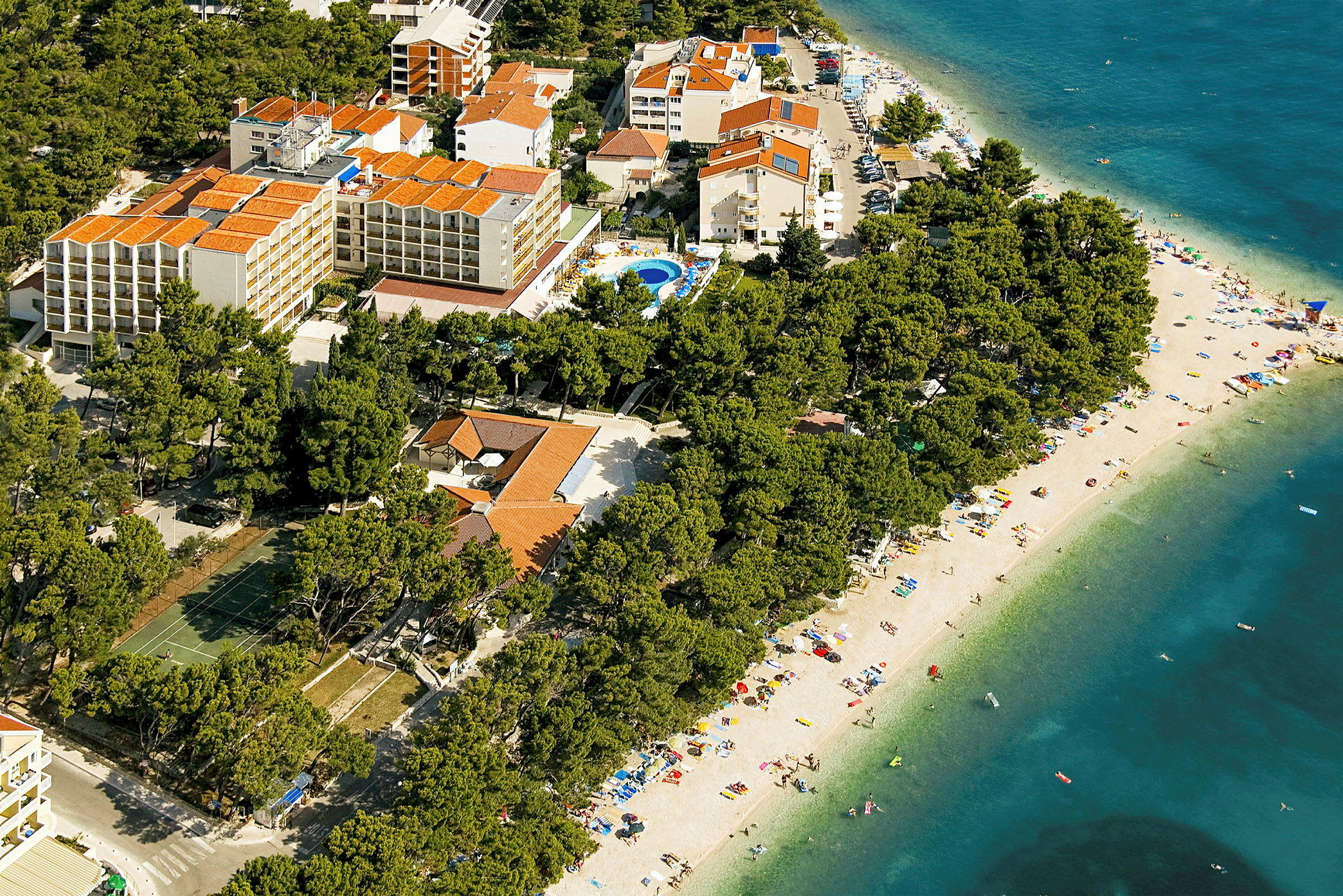 Flygbild som visar hotellbyggnader omgivna av träd med en sandstrand och blått hav i närheten. (AI-genererad text)