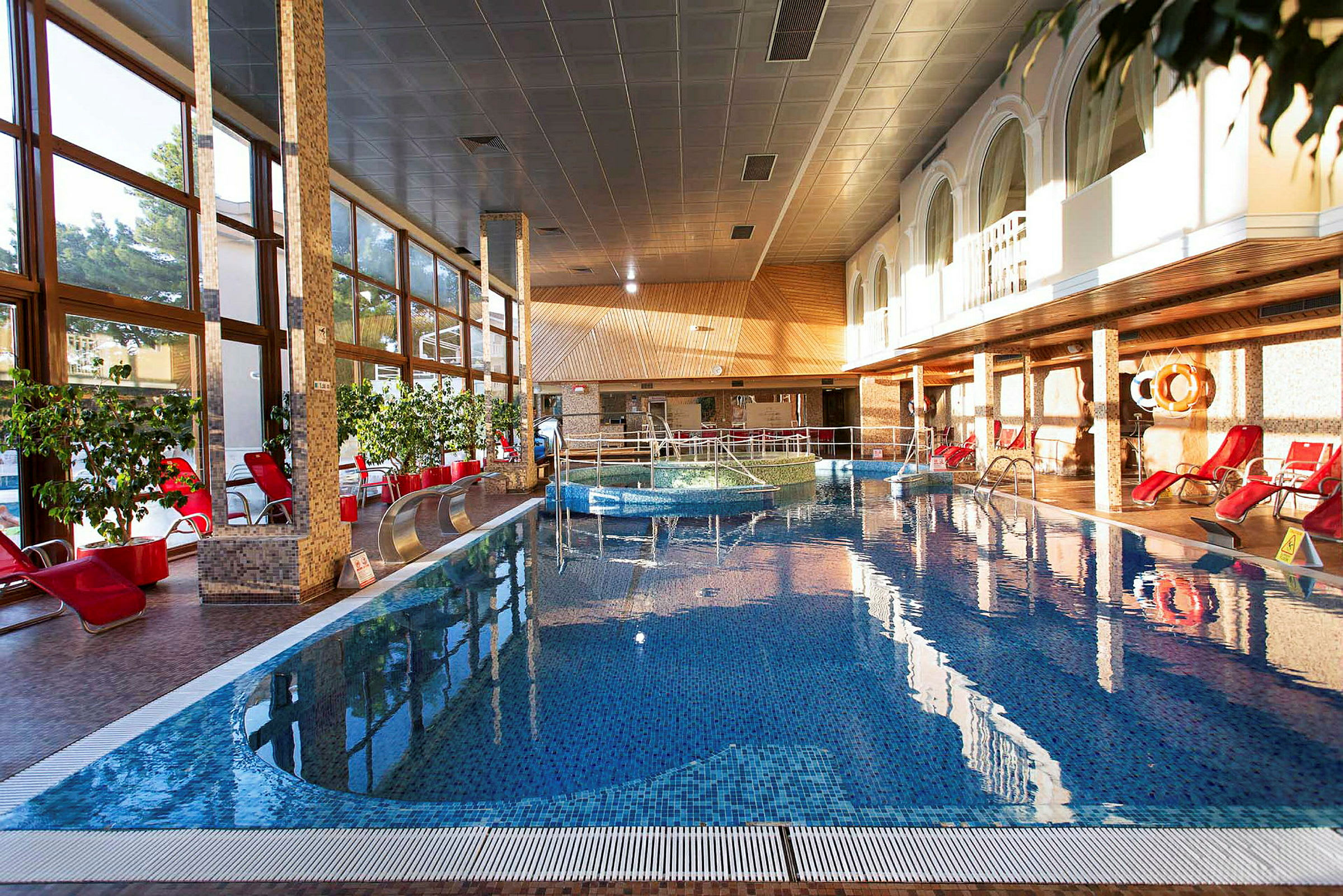 En inomhuspool med röda solstolar runtom, växter och stora fönster. (AI-genererad text)