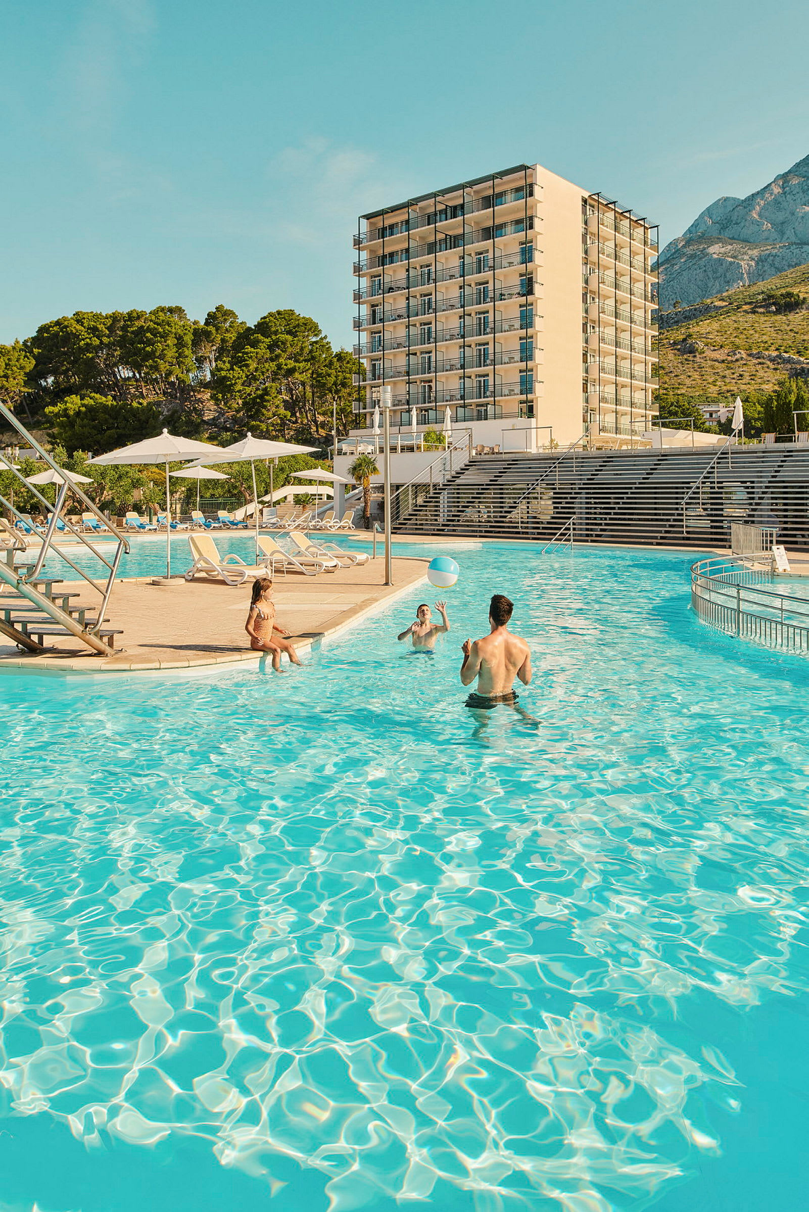 En stor pool framför ett hotell med flera våningar, omgiven av solstolar och parasoller. (AI-genererad text)