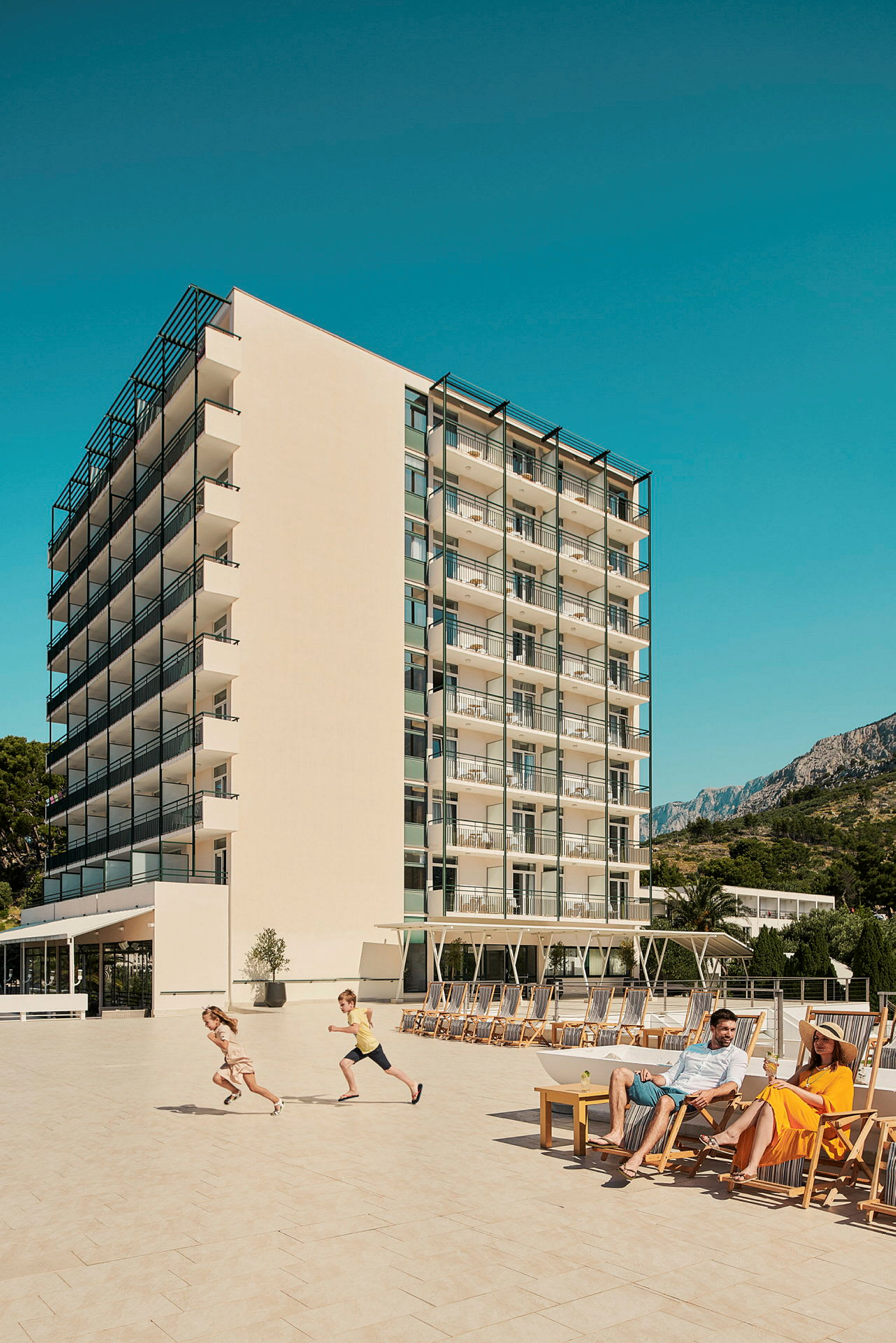 Bild av ett hotell med balkonger i flera våningar, en stor terrass med solstolar, och personer som njuter av solen. (AI-genererad text)