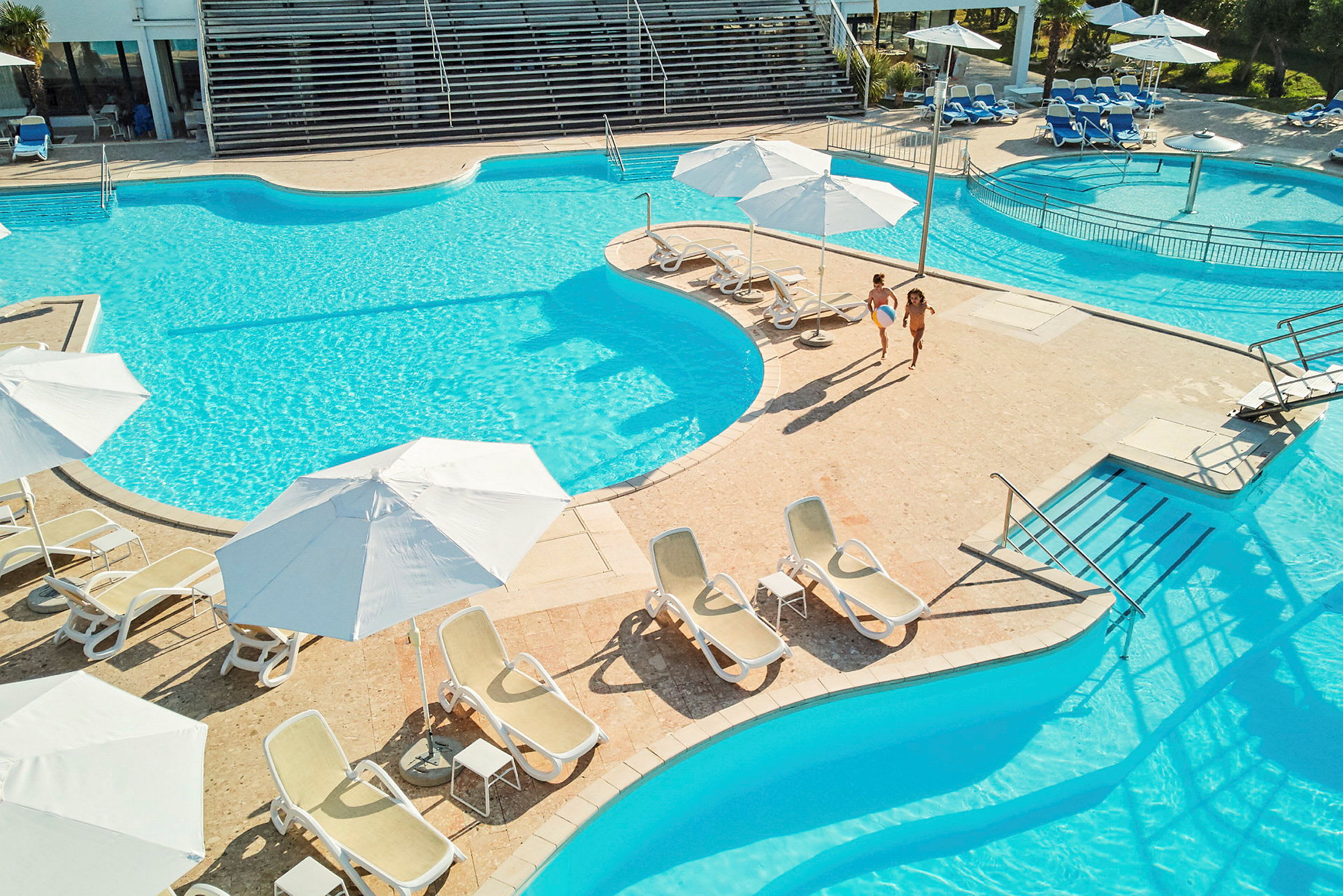 Två personer går vid en pool omgivna av solstolar och parasoller. (AI-genererad text)