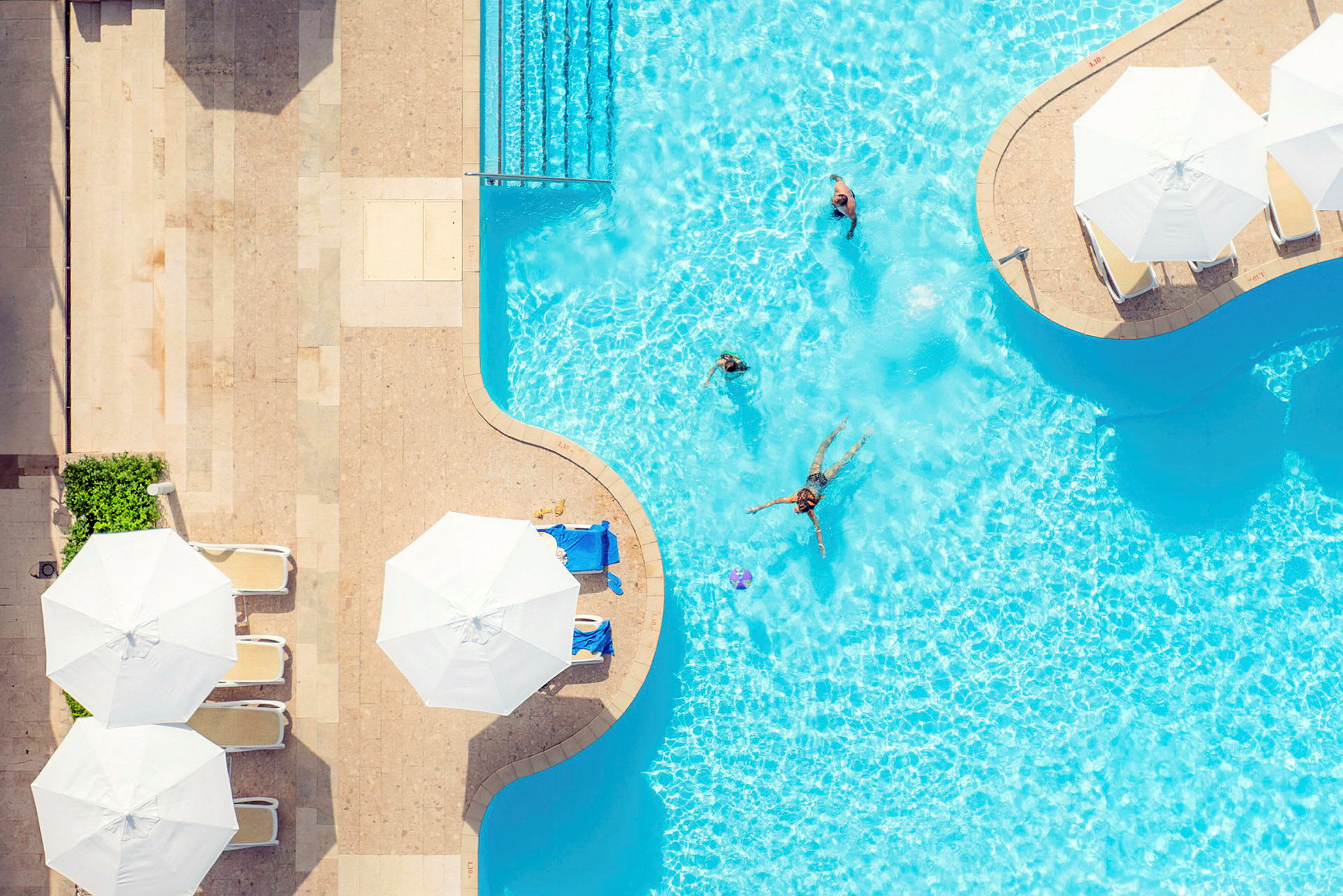 Vy ovanifrån av en pool med några människor som badar och solstolar under parasoller vid poolkanten. (AI-genererad text)