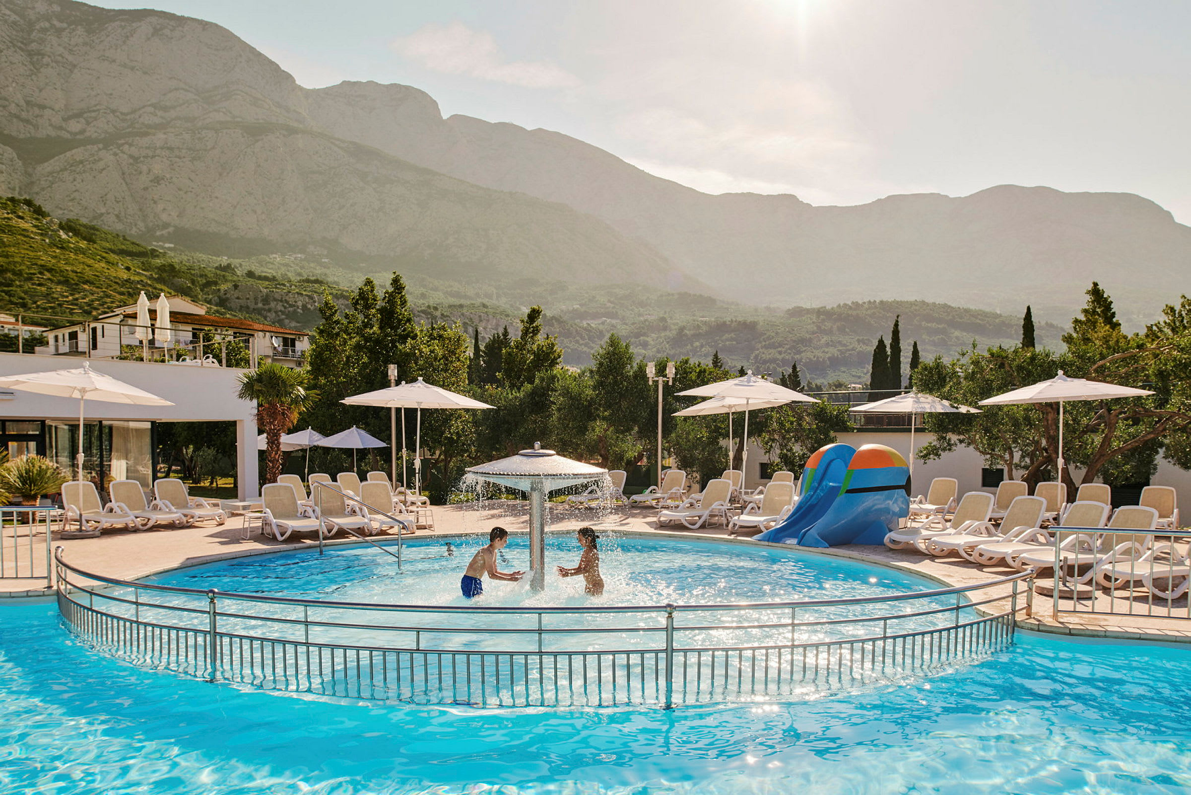 Två barn leker i en pool med vattenfontän och solsängar samt parasoller i bakgrunden. (AI-genererad text)