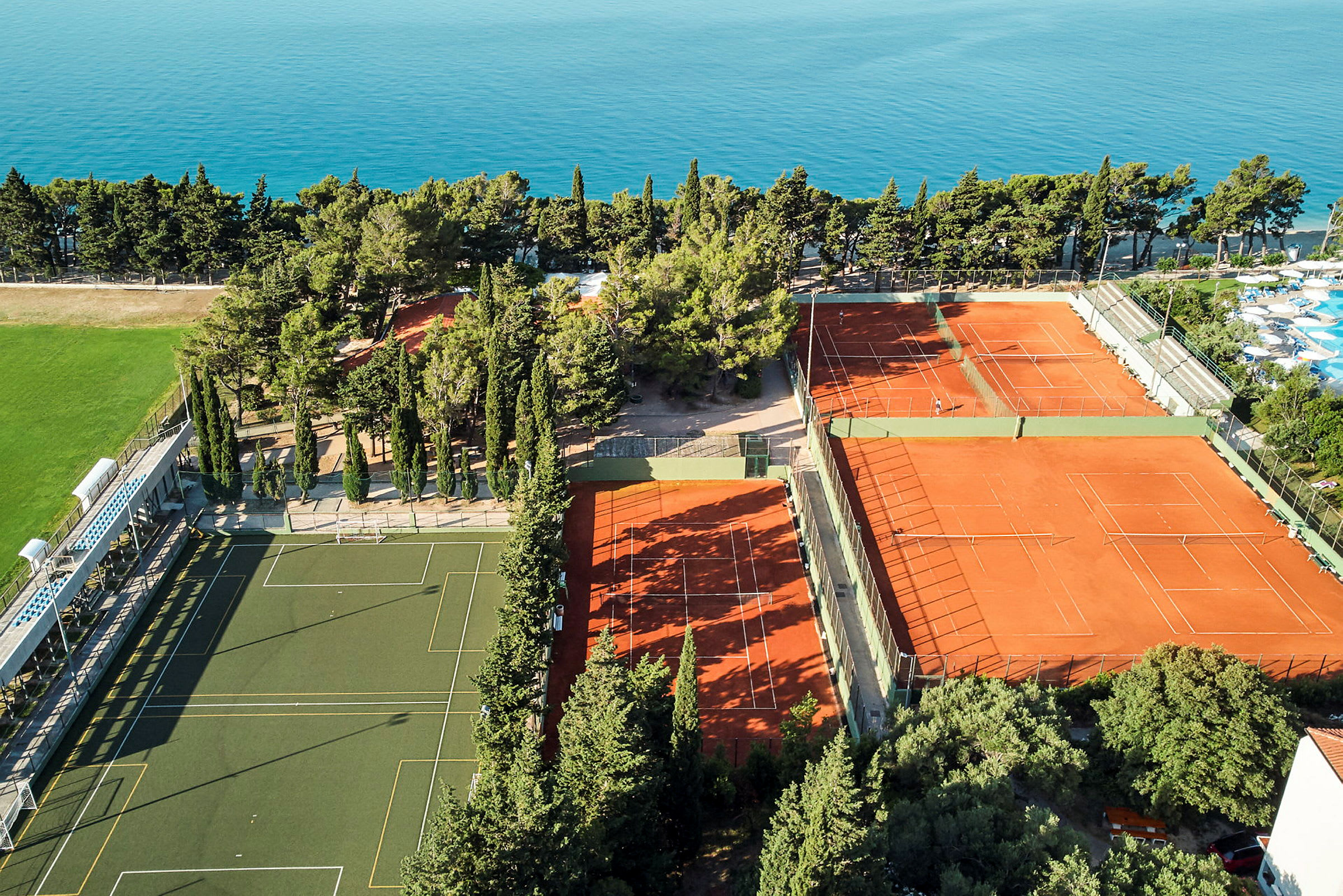 Flygfoto av sportanläggning med tennisbanor och fotbollsplan, omgiven av träd och hav. (AI-genererad text)