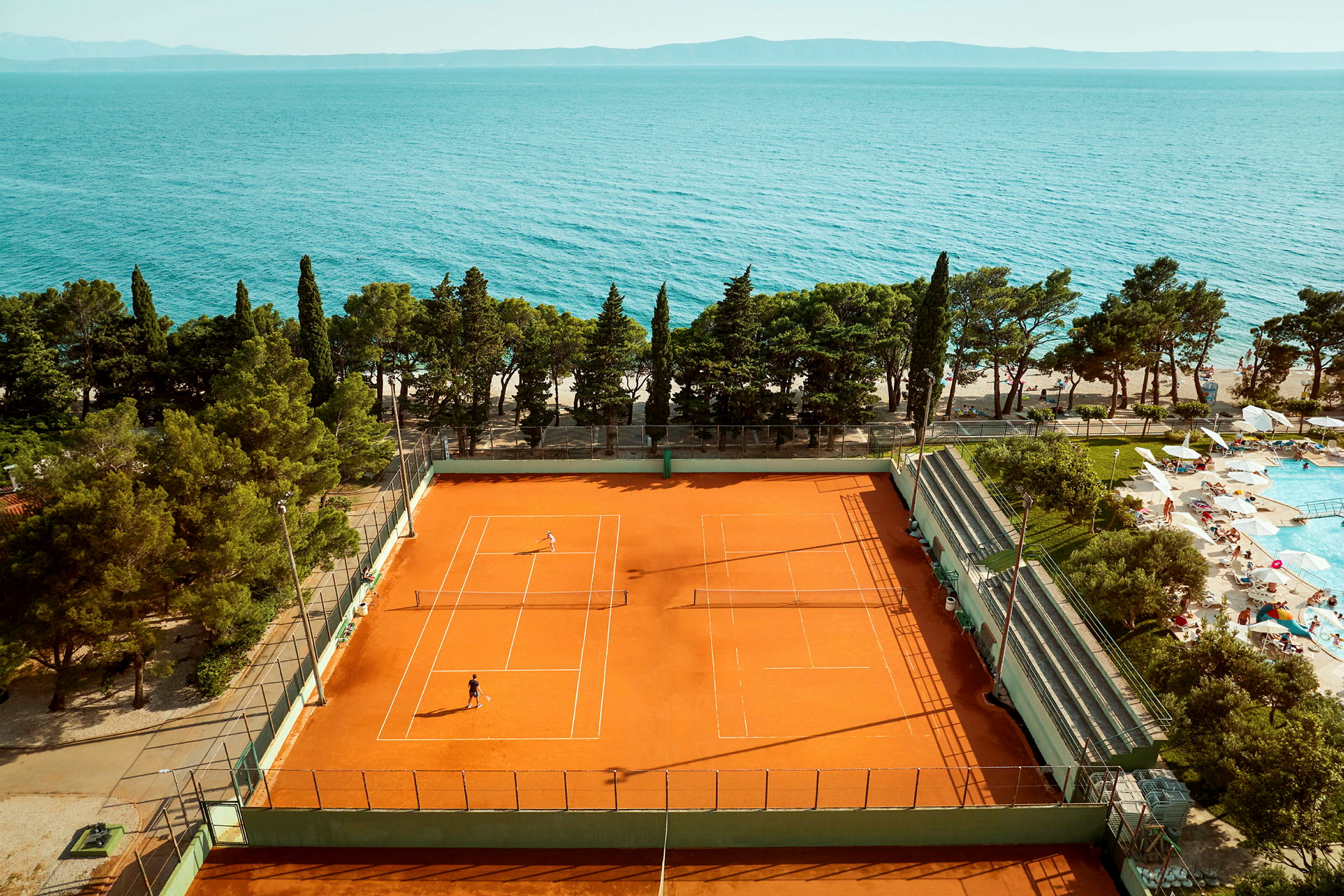 Tennisbana med spelare nära havet och en poolområde. (AI-genererad text)