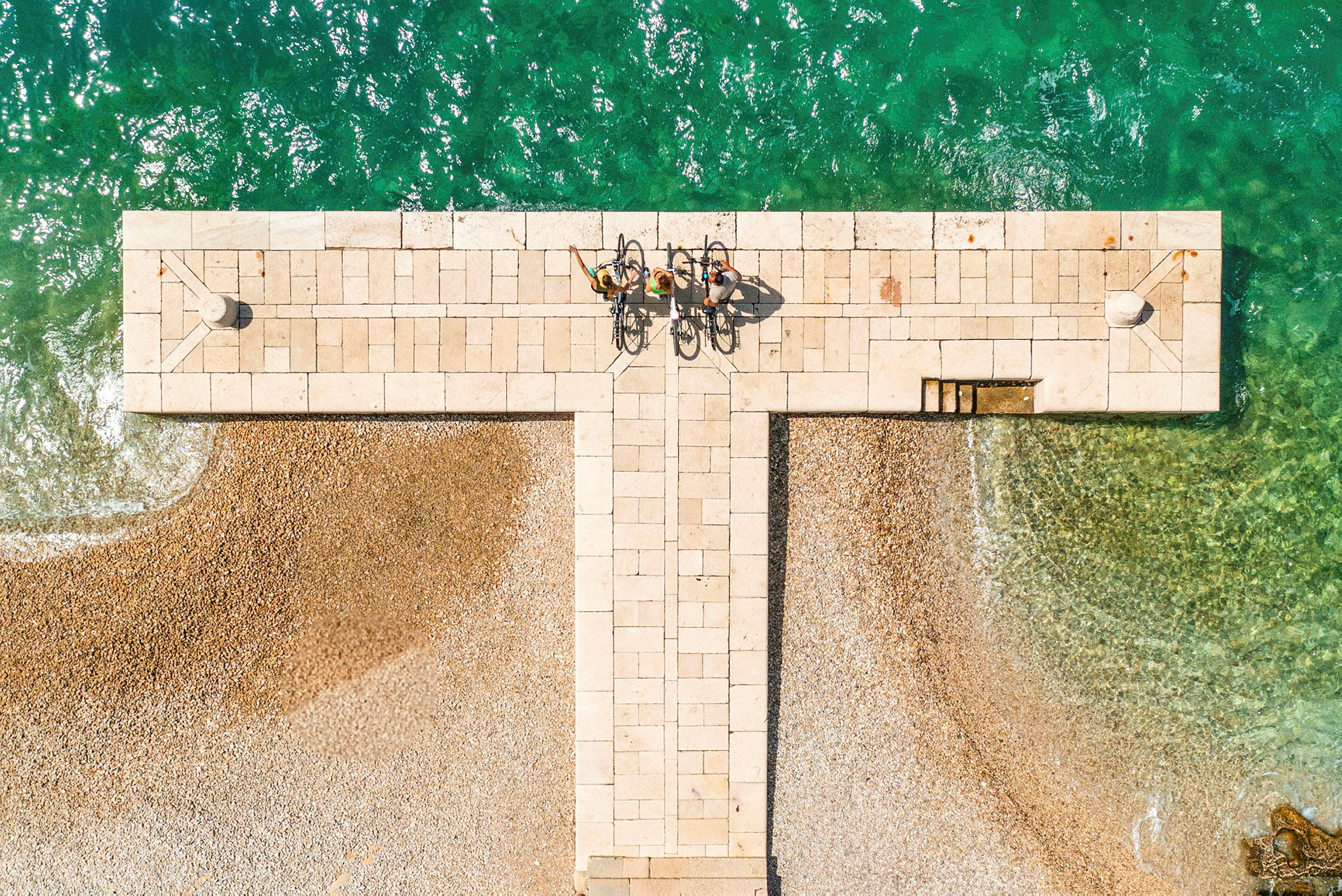 Flygfoto av en stenkaj med tre cyklister nära grönt vatten. (AI-genererad text)