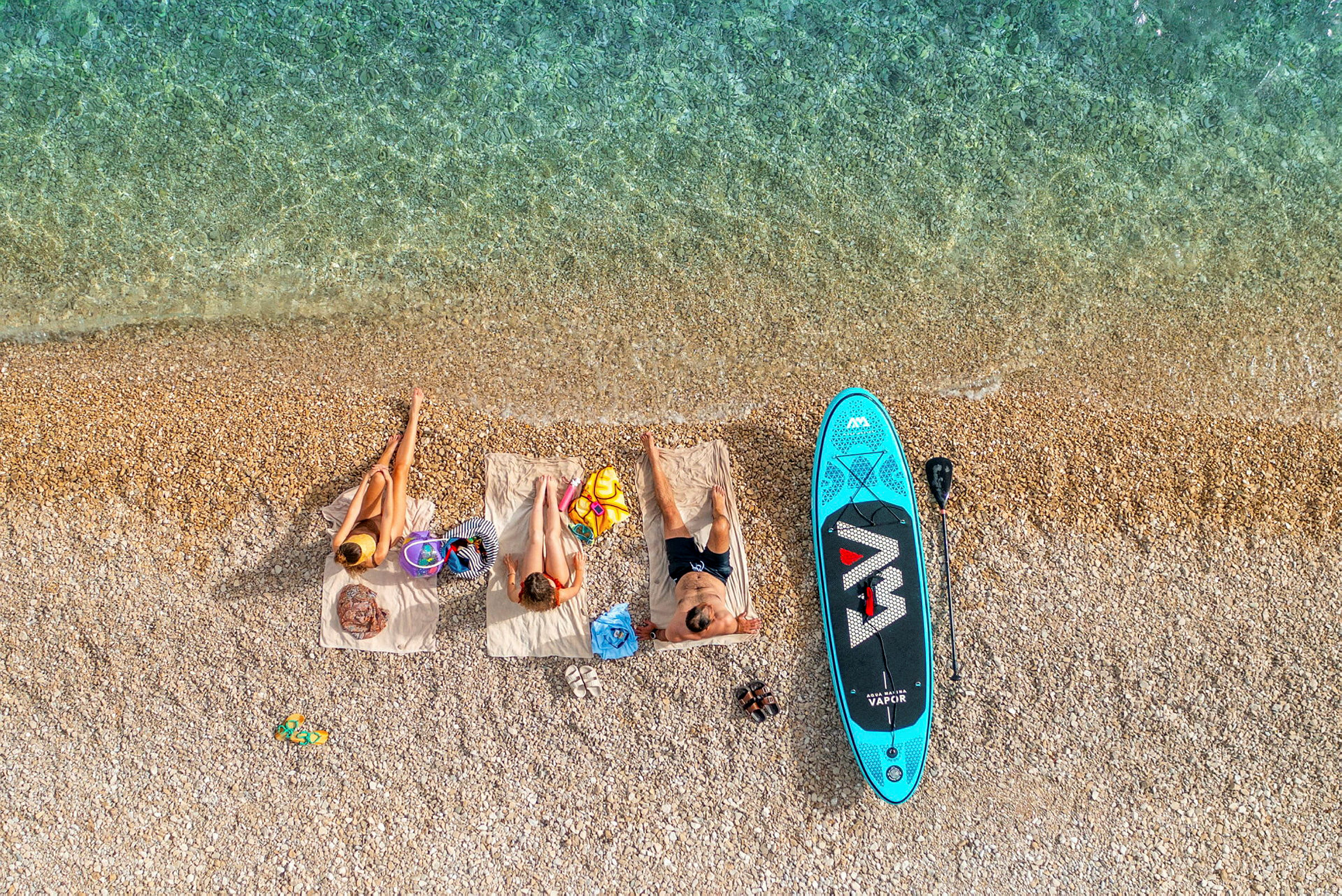 Tre personer som ligger på handdukar vid stranden bredvid en paddleboard. (AI-genererad text)