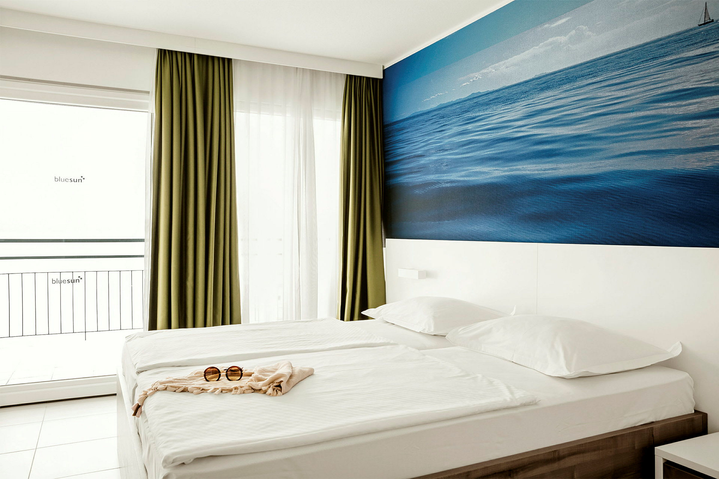 Ett hotellrum med en dubbelsäng, gröna gardiner, en vit vägg med en blå bild av havet och ett par solglasögon på sängen. (AI-genererad text)