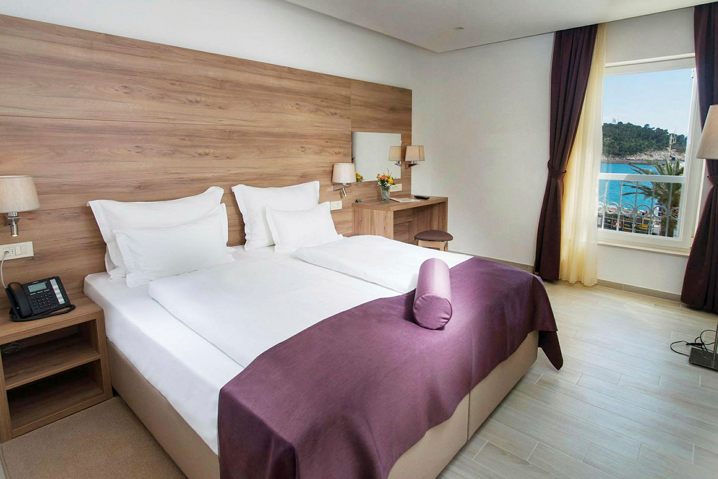 Ett hotellrum med en dubbelsäng, vita lakan och lila överkast. Det finns ett skrivbord med en stol och en spegel. Fönstret visar utsikt mot havet. (AI-genererad text)