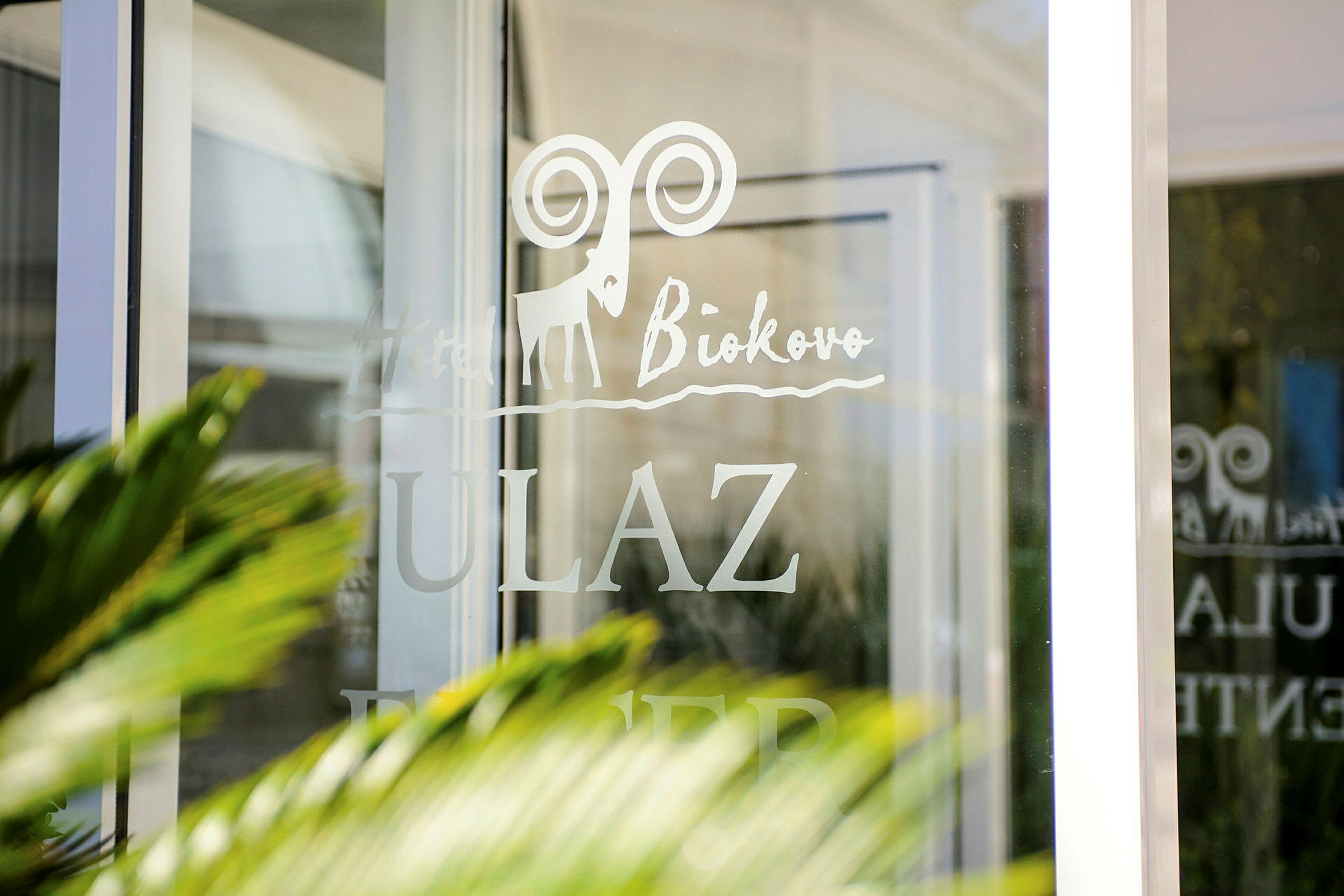 Text på en glasdörr där det står "Hotel Biokovo" och "ULAZ," med en tecknad symbol av en stenbock. (AI-genererad text)