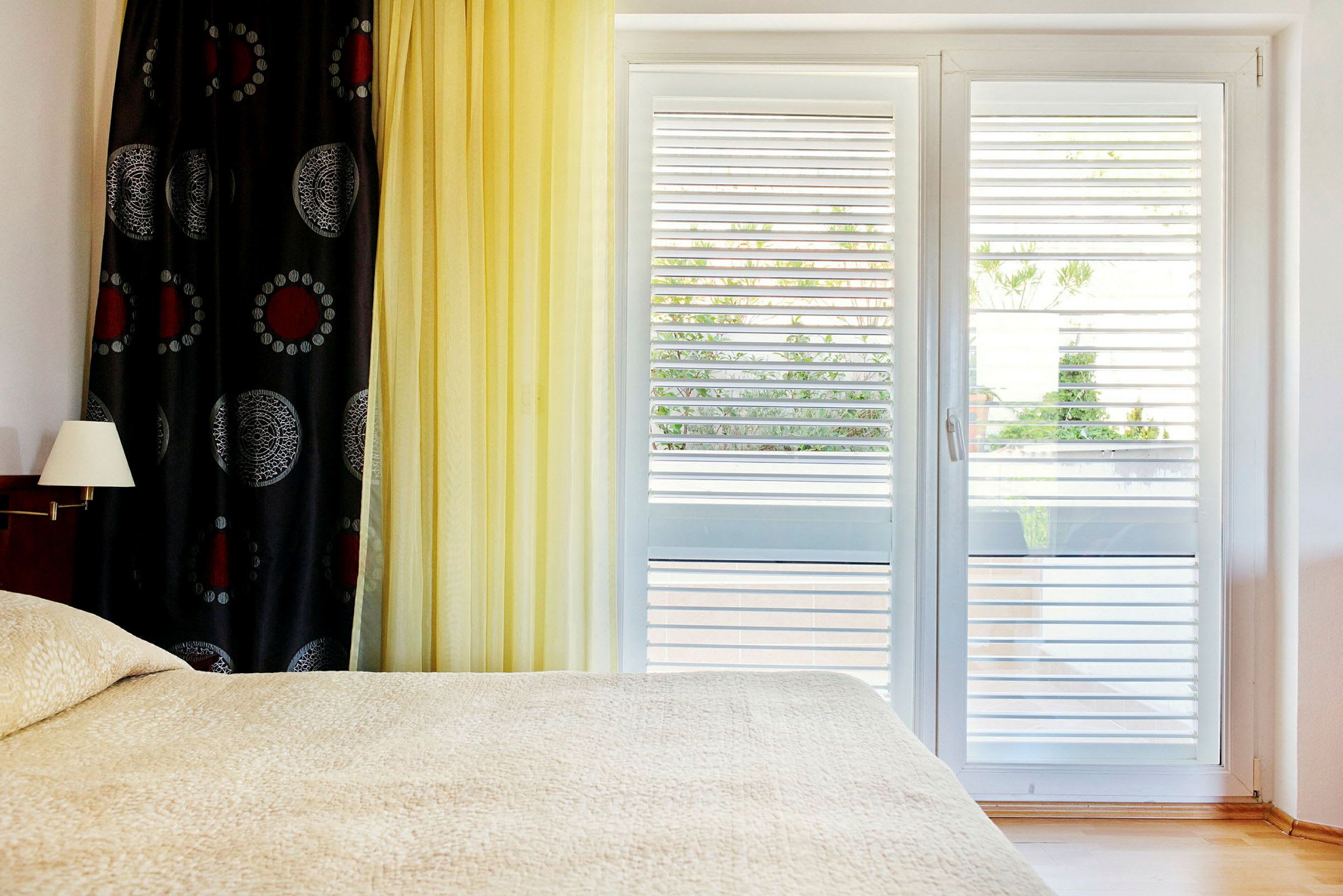 Ett sovrum med en säng, en lampa på väggen och ett fönster med jalusier. (AI-genererad text)