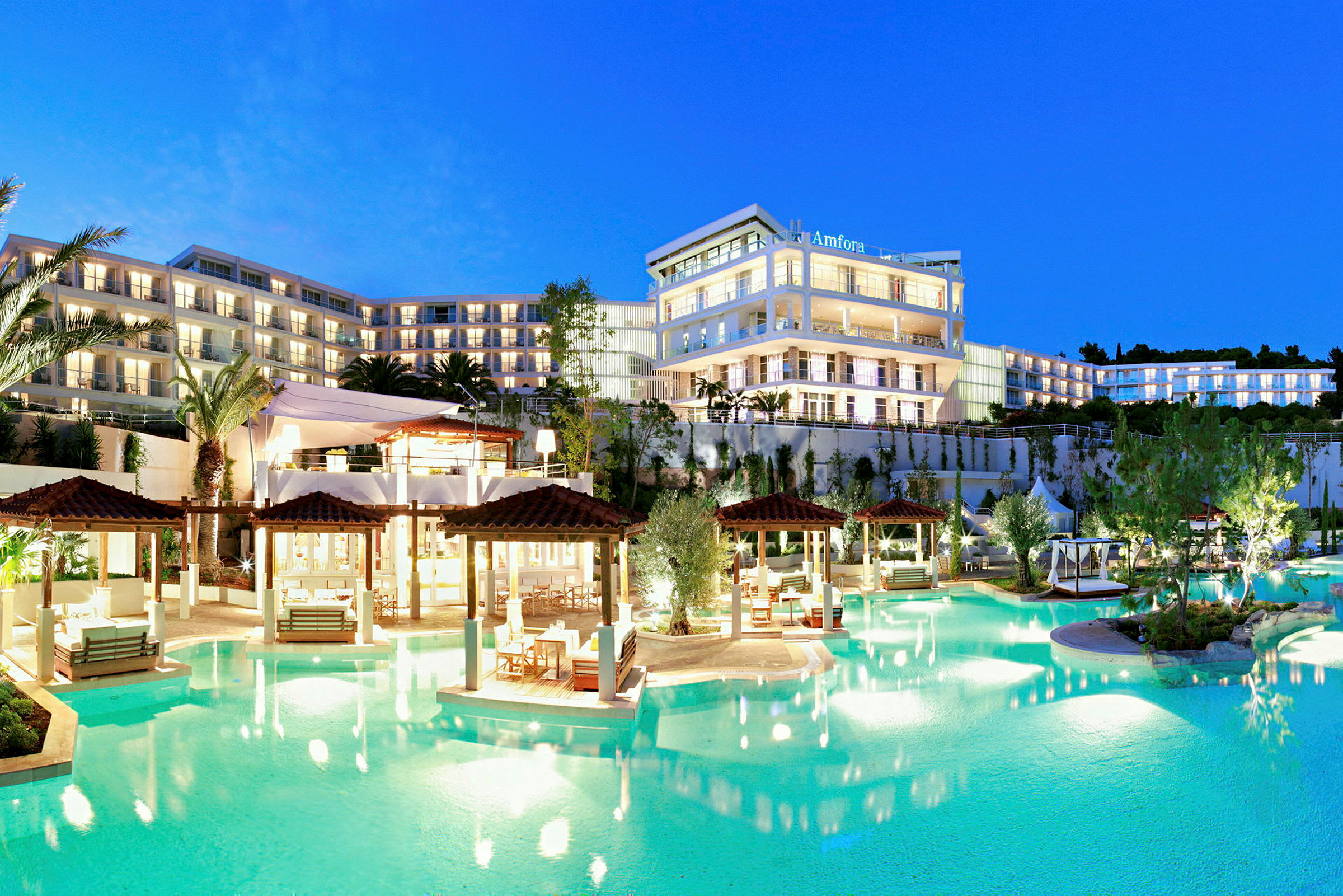 Ett upplyst hotellkomplex med flera våningar vid en pool, omgiven av trädgårdar och palmer, i skymning. (AI-genererad text)