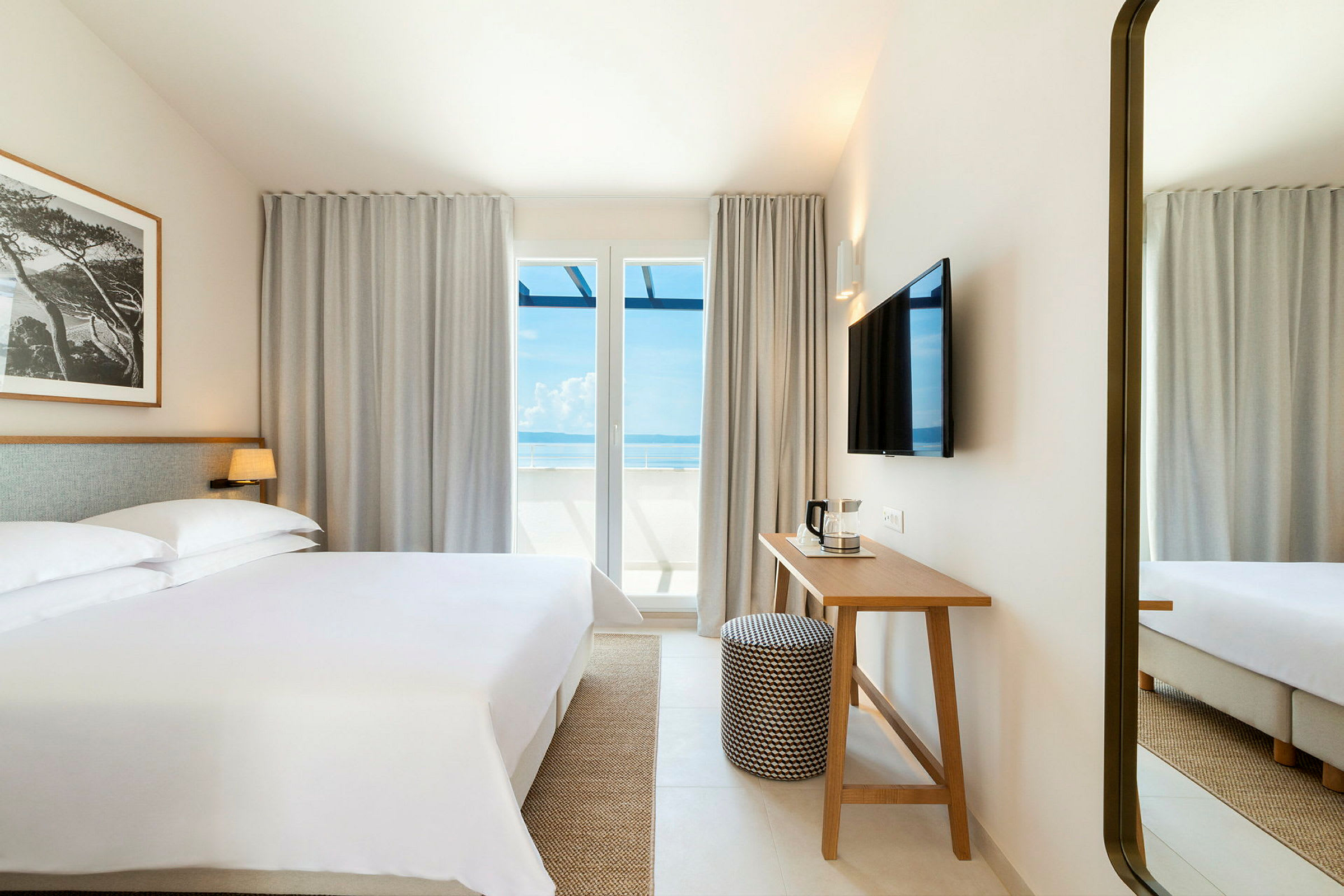 Ett hotellrum med en stor säng, ett skrivbord, och en TV på väggen. Stora fönster med utsikt mot havet. (AI-genererad text)
