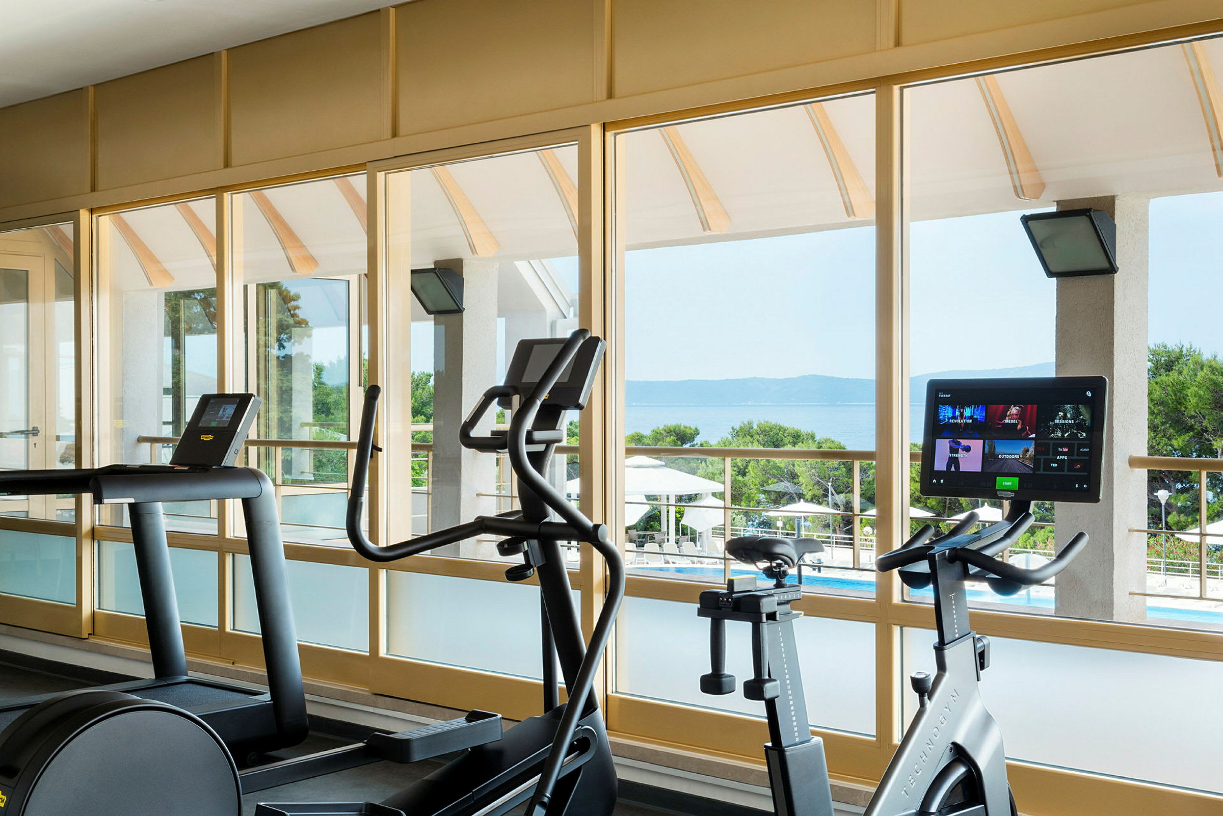 Gym med träningsmaskiner framför stora fönster med utsikt över havet. (AI-genererad text)