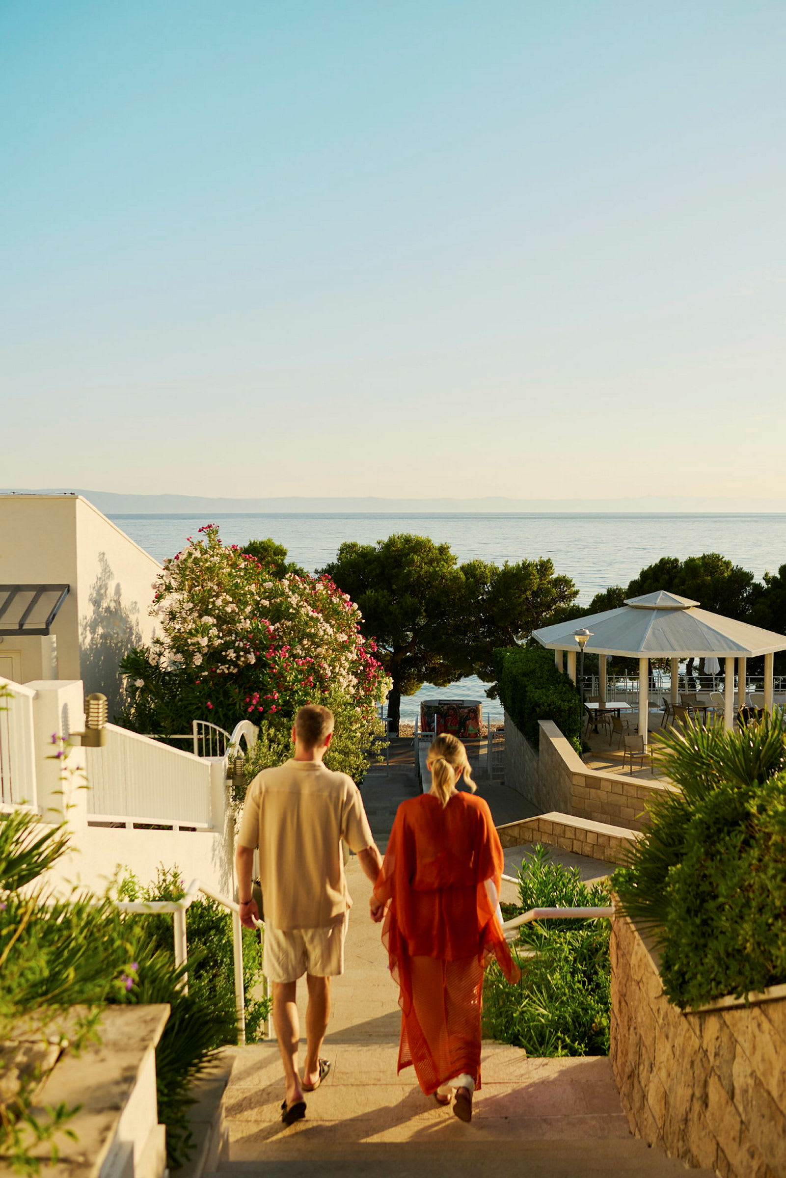 Ett par promenerar nedför en trappa mot havet, med grönska och byggnader omkring. (AI-genererad text)