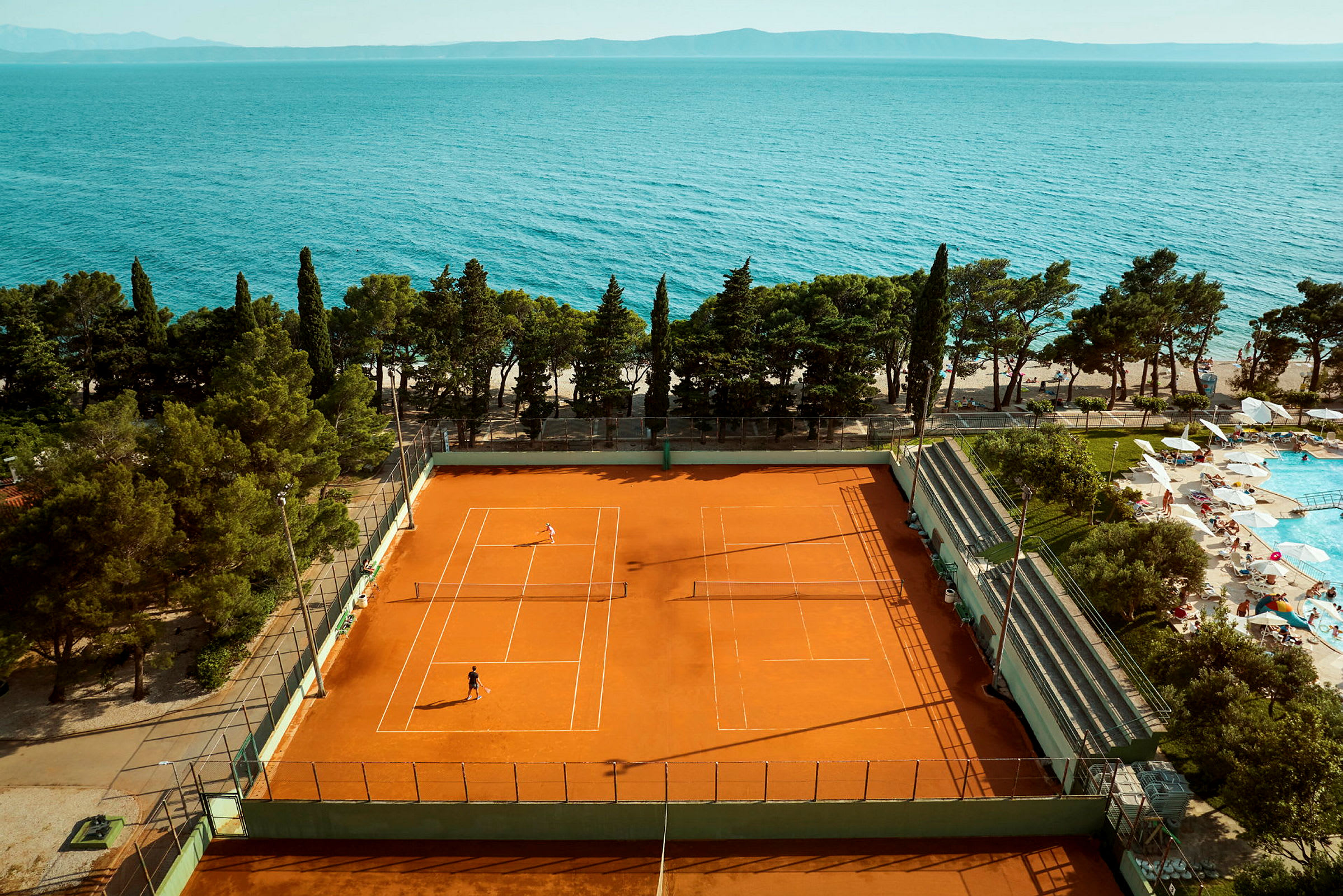 En tennisplan med spelare och en poolområde med solstolar bredvid, samt hav i bakgrunden. (AI-genererad text)