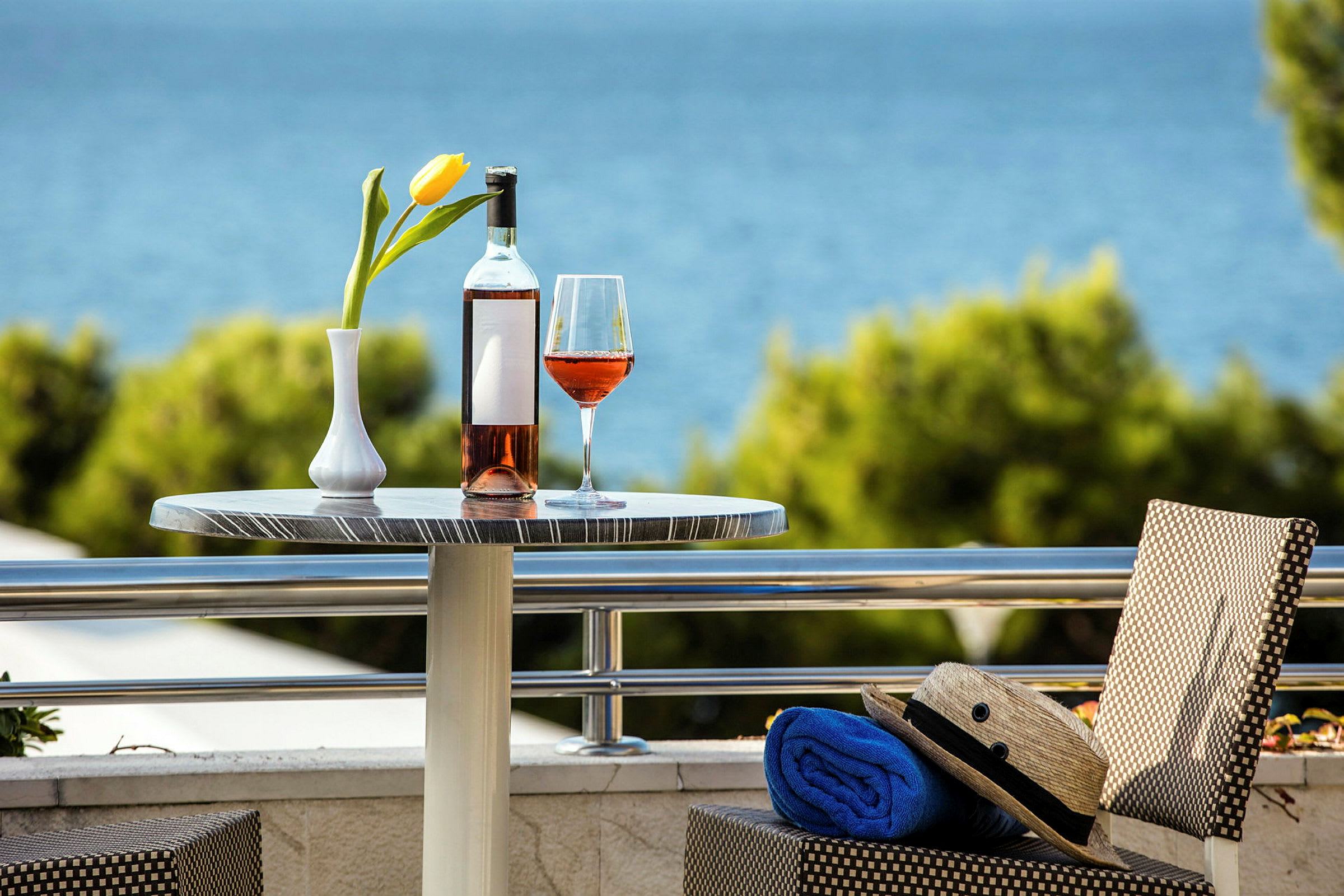 Ett bord med en flaska och ett glas rosévin samt en gul tulpan i vas, med en utsikt över havet i bakgrunden. (AI-genererad text)