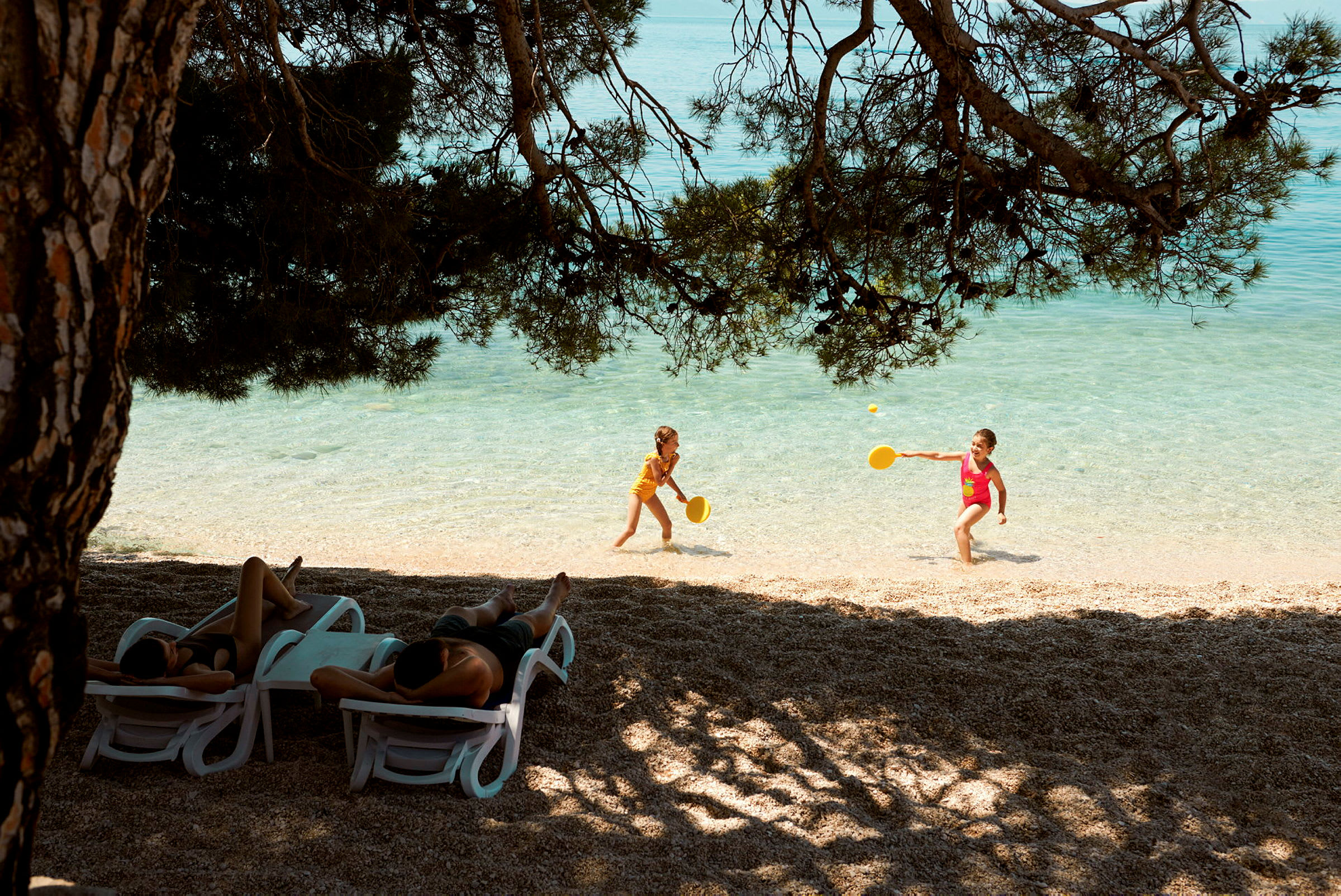 Två barn leker med racketar i vattenbrynet på en strand. Flera personer ligger på solstolar under skuggande träd. (AI-genererad text)