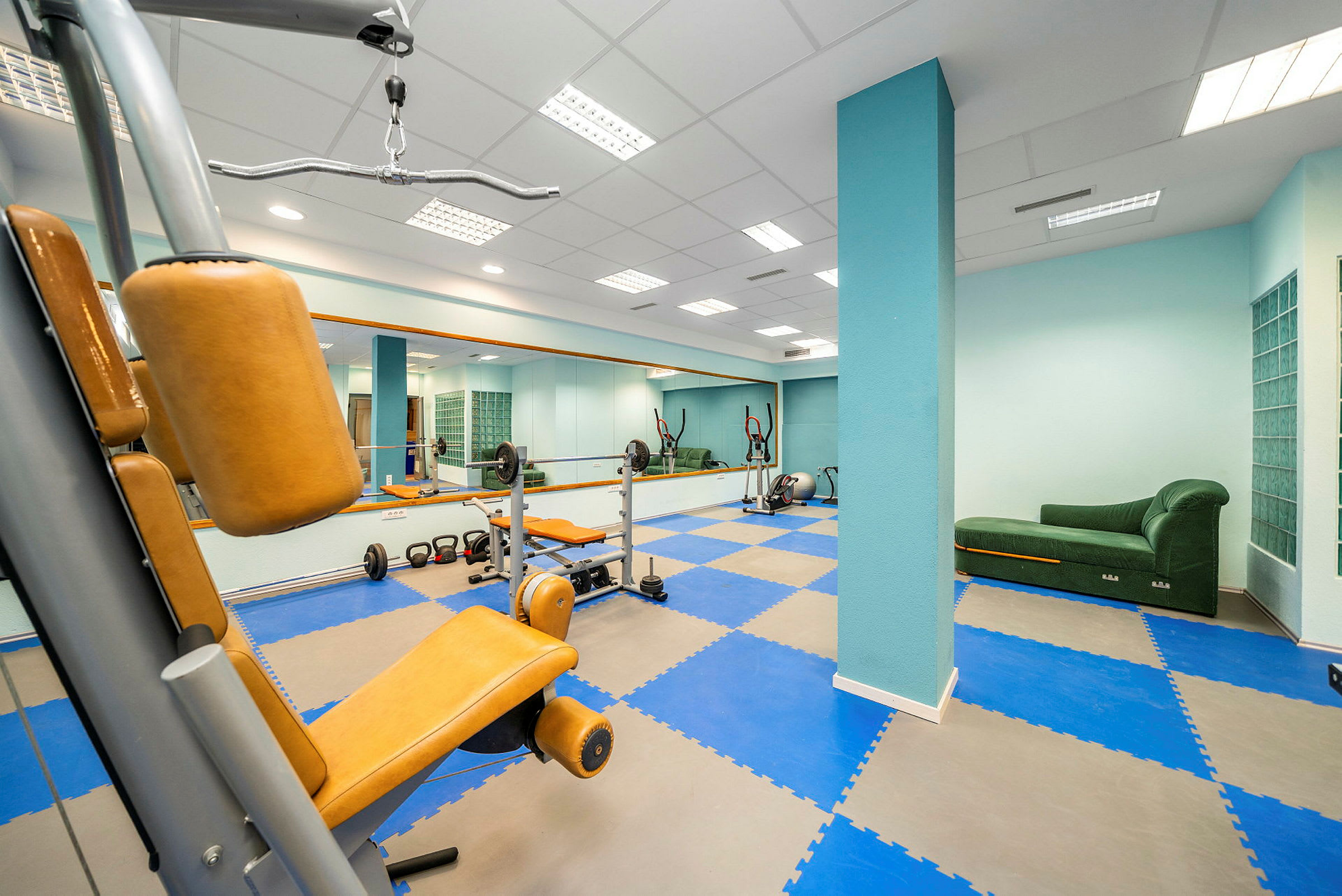 Ett gym med träningsmaskiner, fria vikter, och en grön soffa. (AI-genererad text)