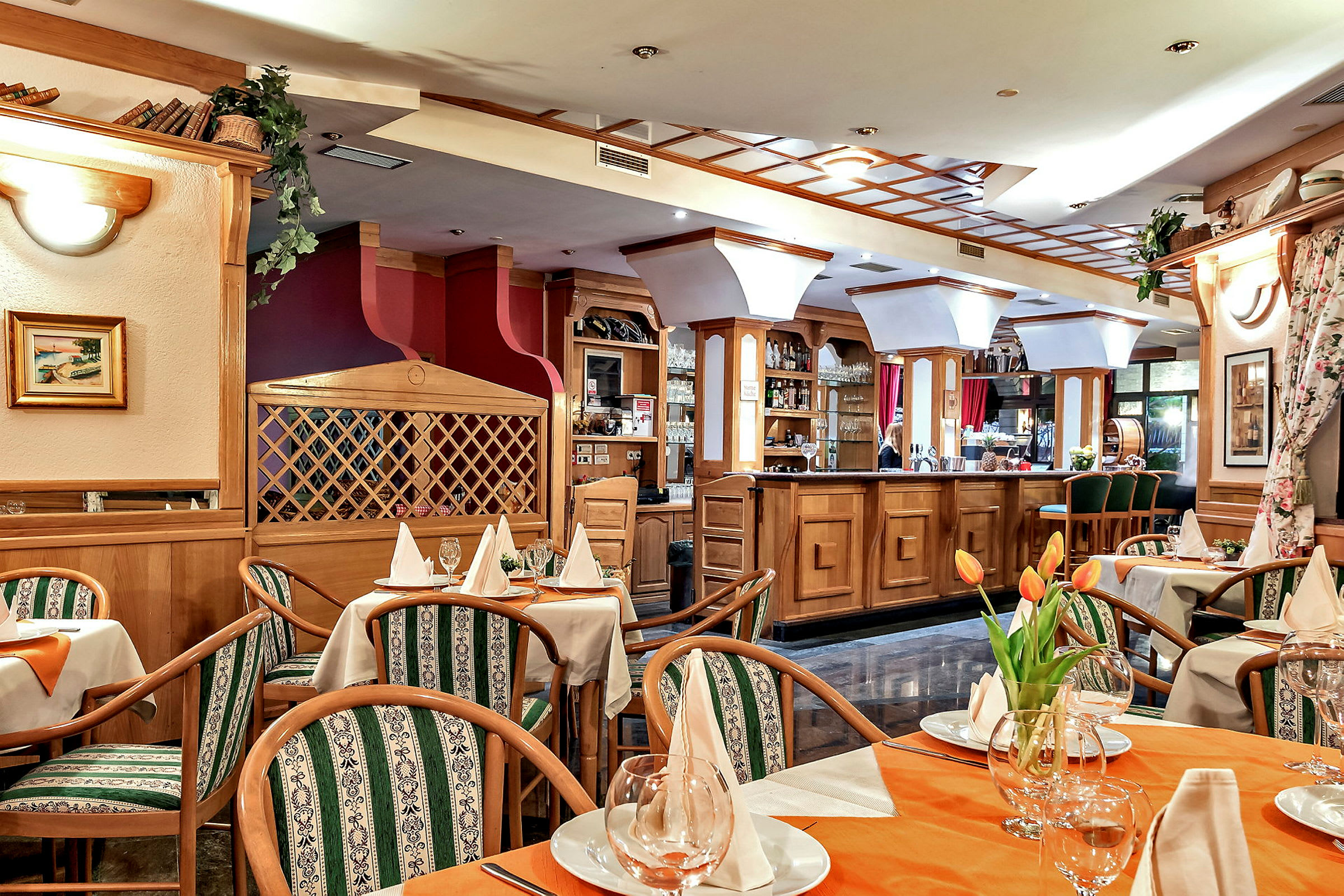En inredd restaurang med bord som är dukade med vita servetter och glas. Stolar med gröna mönstrade dynor omger borden, och en bardisk i trä är synlig i bakgrunden. (AI-genererad text)