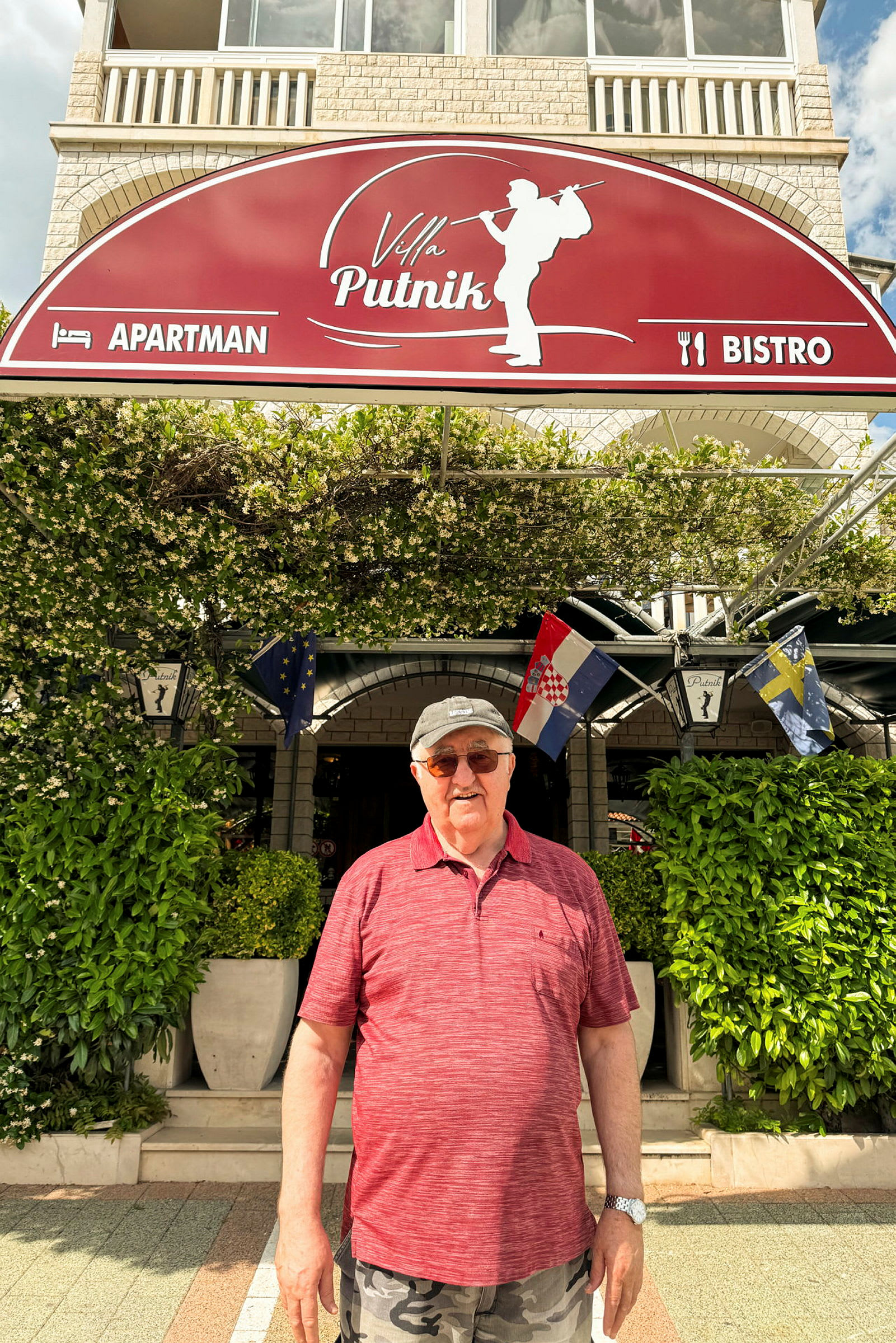 Bild på en person stående framför en byggnad med en skylt där det står "Villa Putnik" och som är omgiven av gröna växter. (AI-genererad text)